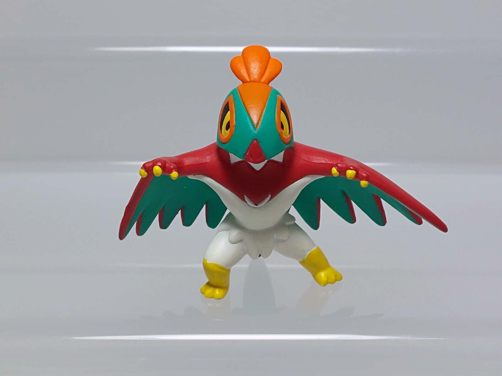 Hawlucha 直視前方 壁紙 — 一個色彩繽紛的塑膠玩具公仔，外形像鳥類的寵物小精靈，帶有紅色、綠色和黃色的特徵，站在反光的白色表面上。