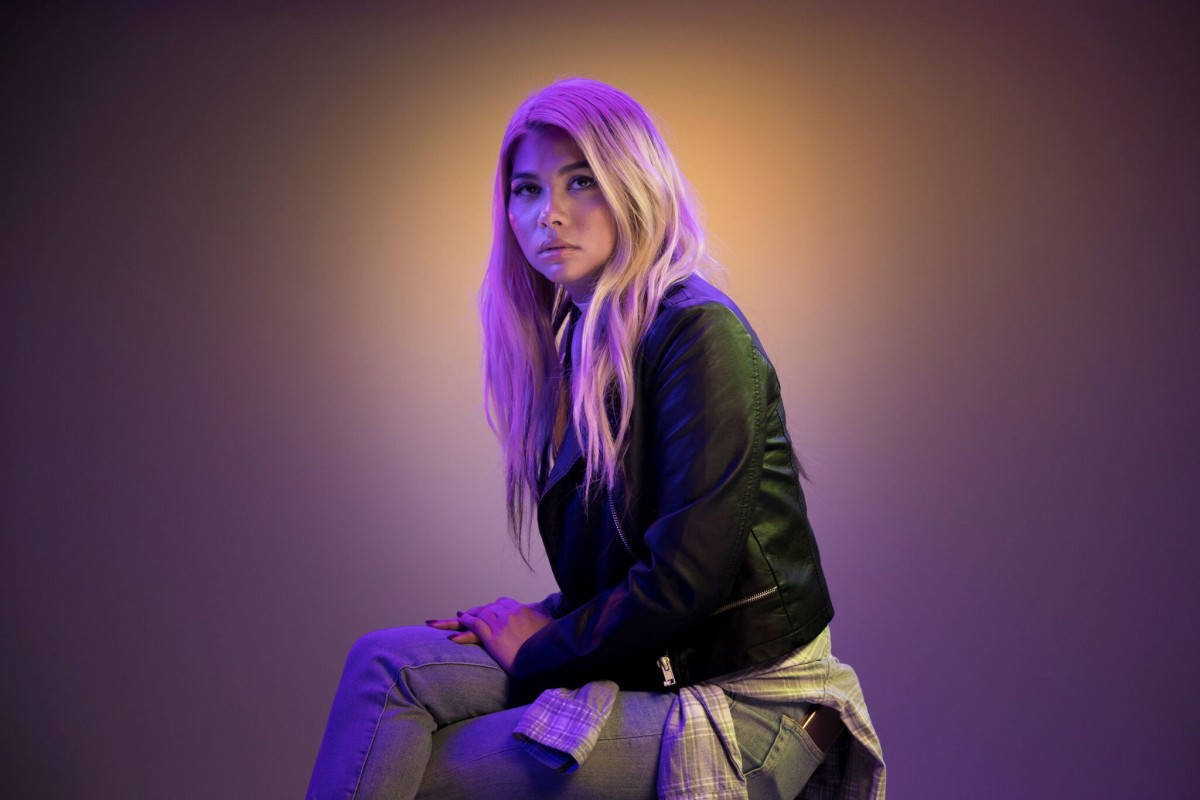 Hayley Kiyoko 皮衣拍攝造型 壁紙 — 一位年輕女性留著金色長髮，坐在充滿紫色和橙色燈光的攝影棚中，身穿黑色皮夾克和牛仔褲。