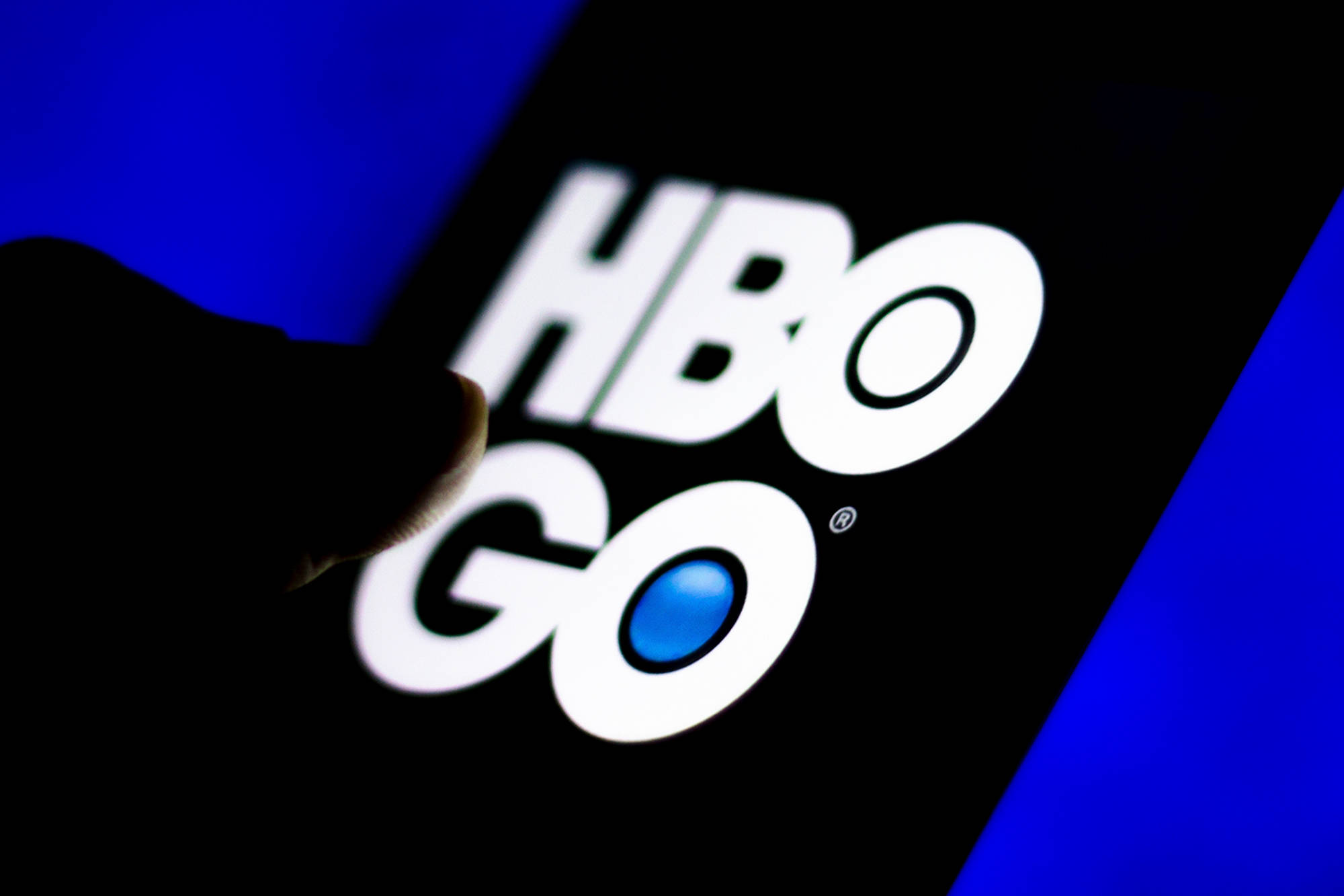 HBO Go 標誌 壁紙 — 手指在深藍色背景上輕觸智能手機屏幕上發光的HBO GO標誌。