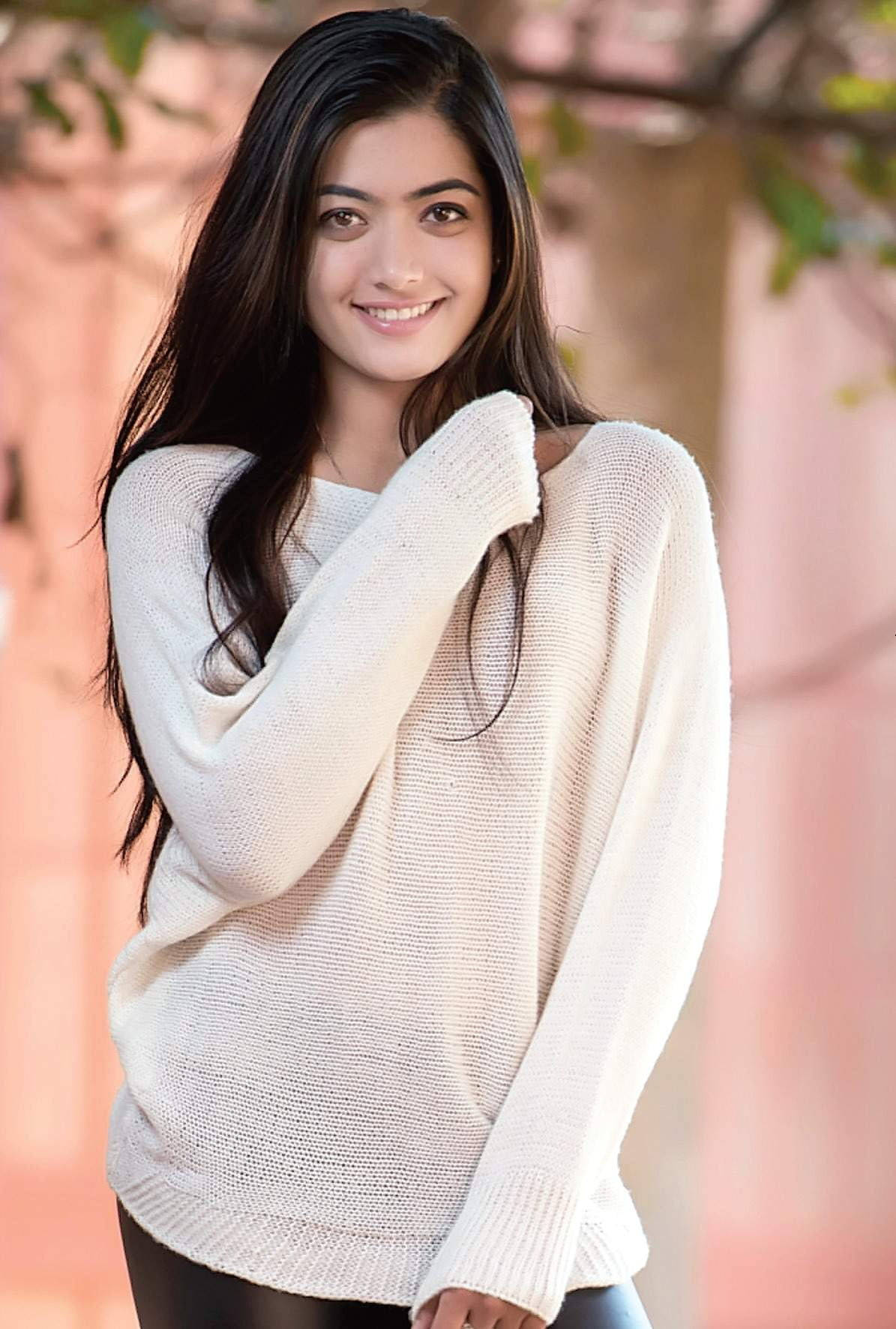簡約拉什米卡·曼丹納 HD 壁紙 — 一位年輕女性留著長長的黑髮，穿著舒適的露肩奶油色毛衣，在戶外溫暖地微笑。