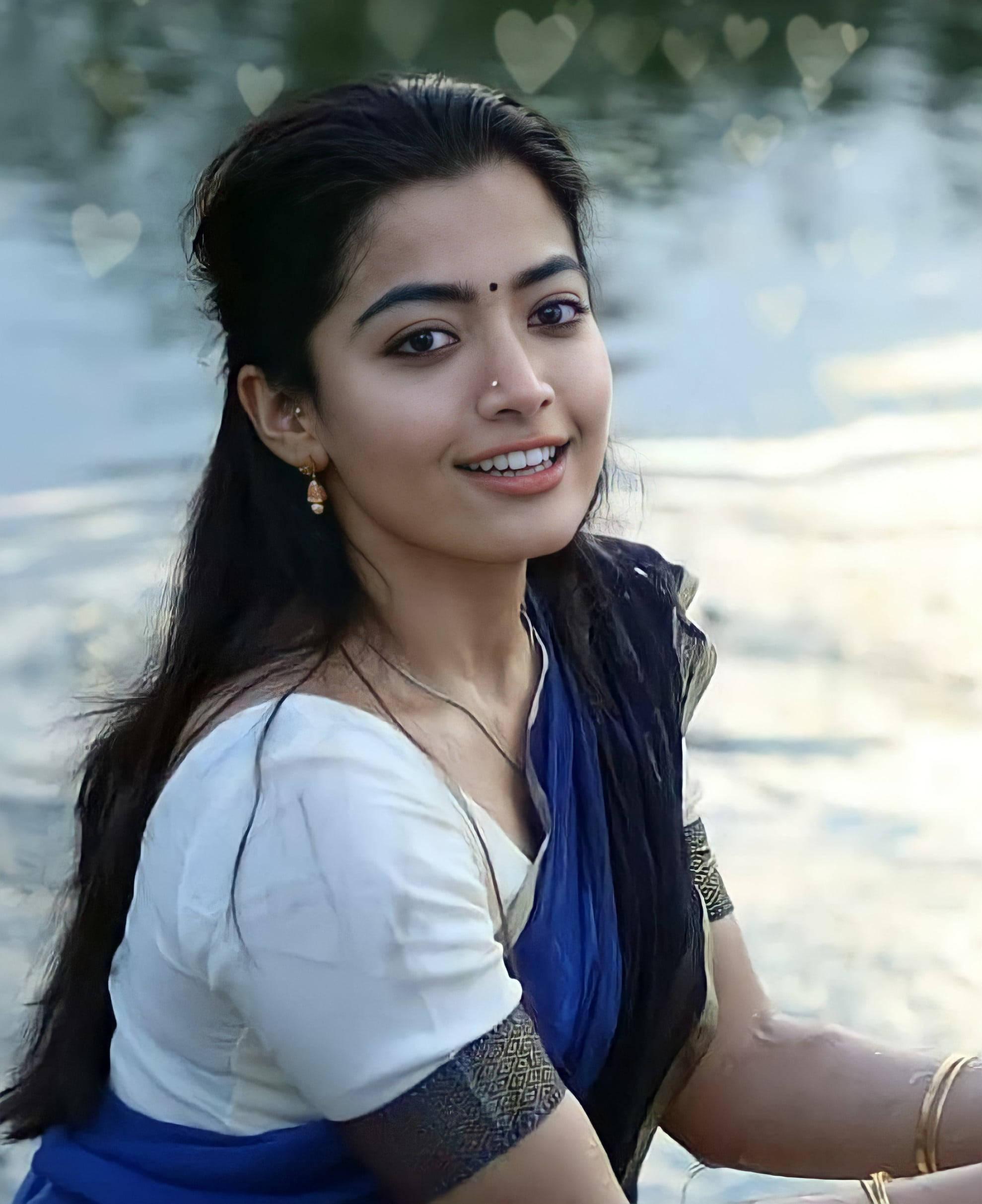 拉什米卡·曼丹納 HD 鼻環特寫 壁紙 — 一位微笑的女子身穿傳統藍白服飾，坐在水邊，背景有心形散景。