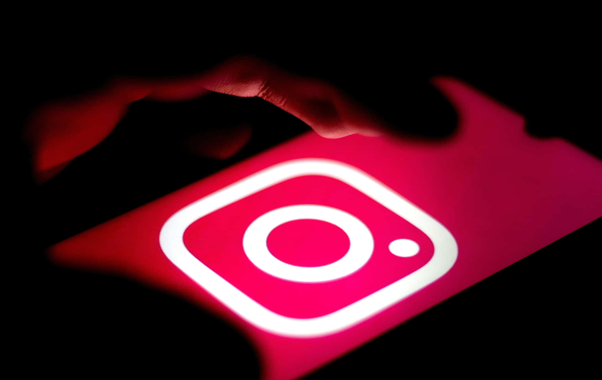  壁紙 — 黑暗中，一隻手懸浮在顯示著發光紅色Instagram標誌的手機屏幕上。