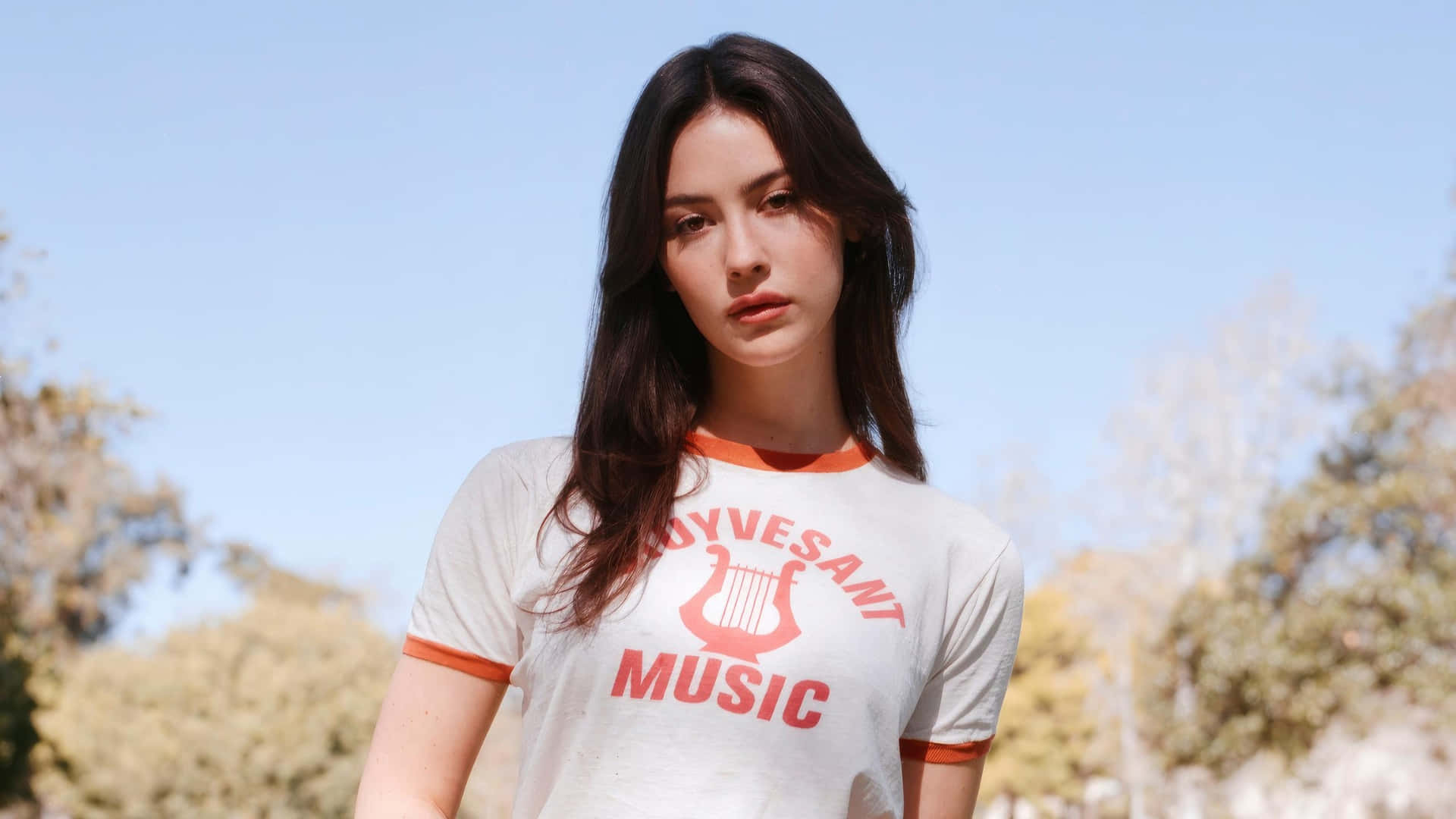  壁紙 — 一位黑髮年輕女性穿著白色T恤，帶有橙色飾邊和紅色 'Hoyvesant Music' 文字及豎琴圖案，背景是晴朗藍天和模糊的樹木。