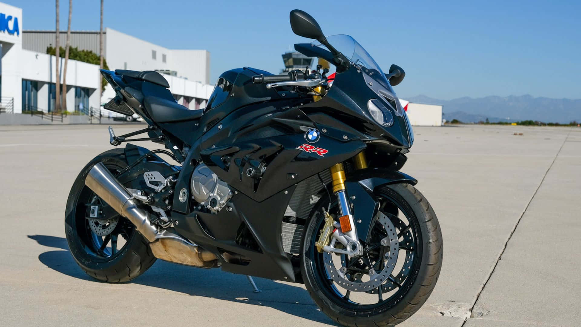  壁紙 — 一輛型格黑色BMW R1200RR運動電單車在陽光下閃耀，停泊在混凝土停機坪上，背景有遠山和管制塔。