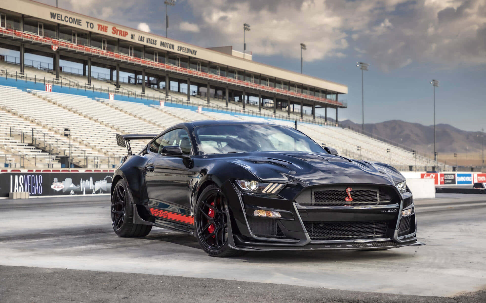  壁紙 — 一架帶有紅色點綴的時尚黑色福特野馬 Shelby GT500 在陰天天空下，停泊在拉斯維加斯賽車場的賽道上。