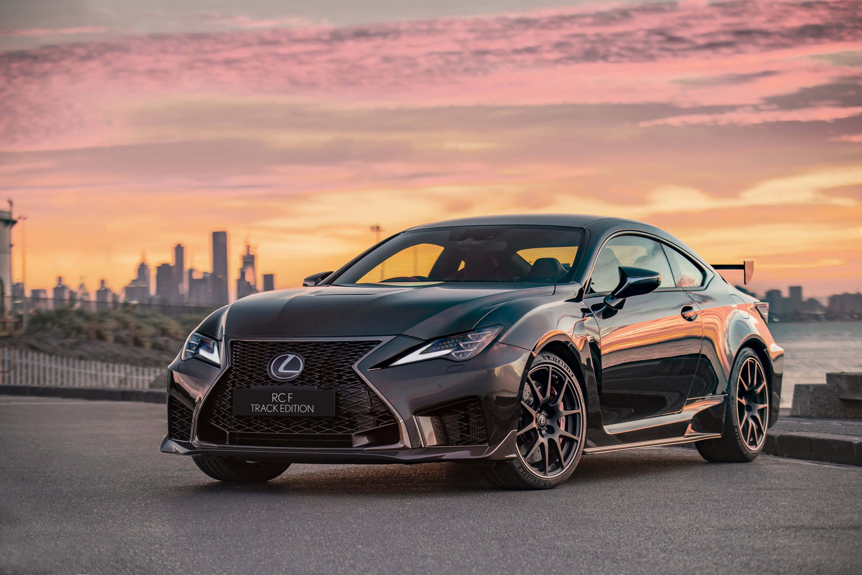 Lexus Rc F 桌布 壁紙 — 一輛時尚的黑色Lexus RC F Track Edition在充滿活力的粉紅色和橙色日落及城市天際線背景下閃閃發光。