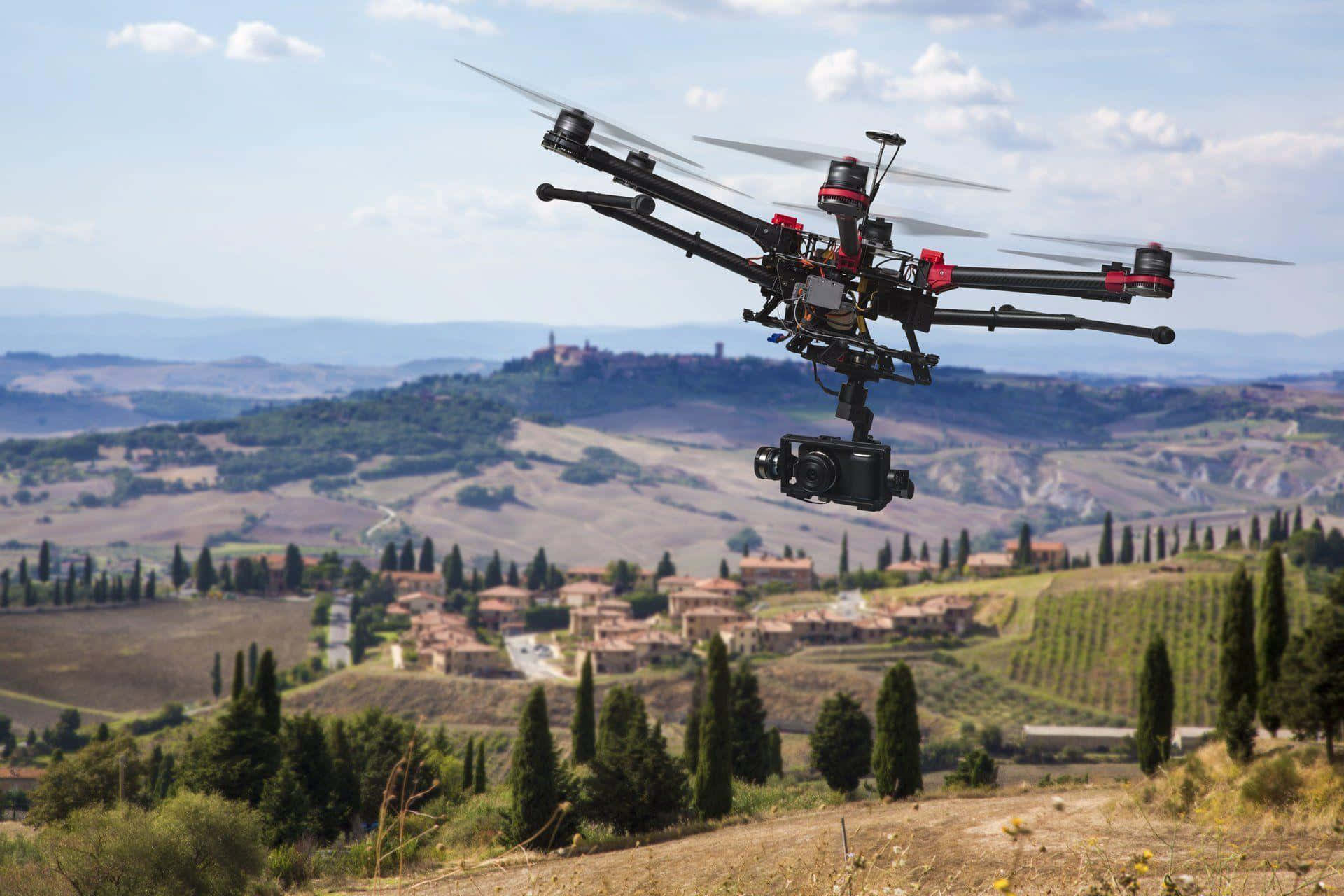 Drone Camera 桌布 壁紙 — 一架帶有紅色點綴的黑色無人機在陽光明媚的托斯卡納鄉村上空盤旋，下方是起伏的丘陵、葡萄園和遠處山頂上的村莊，天空湛藍，白雲點點。