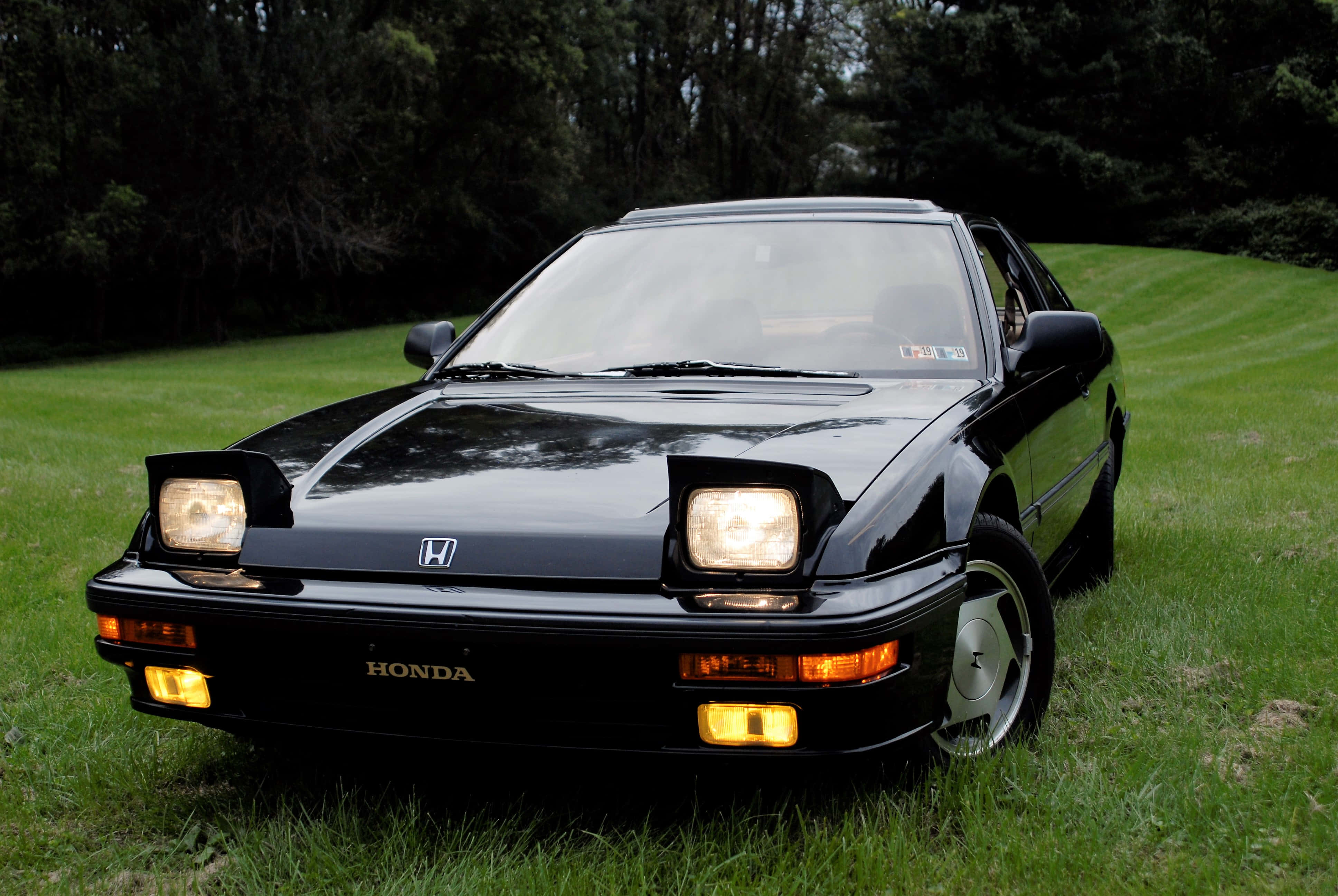 Honda Prelude 桌布 壁紙 — 一輛時尚的黑色本田跑車，車頭燈閃閃發光，停泊在茂密樹木環繞的翠綠草坪上。