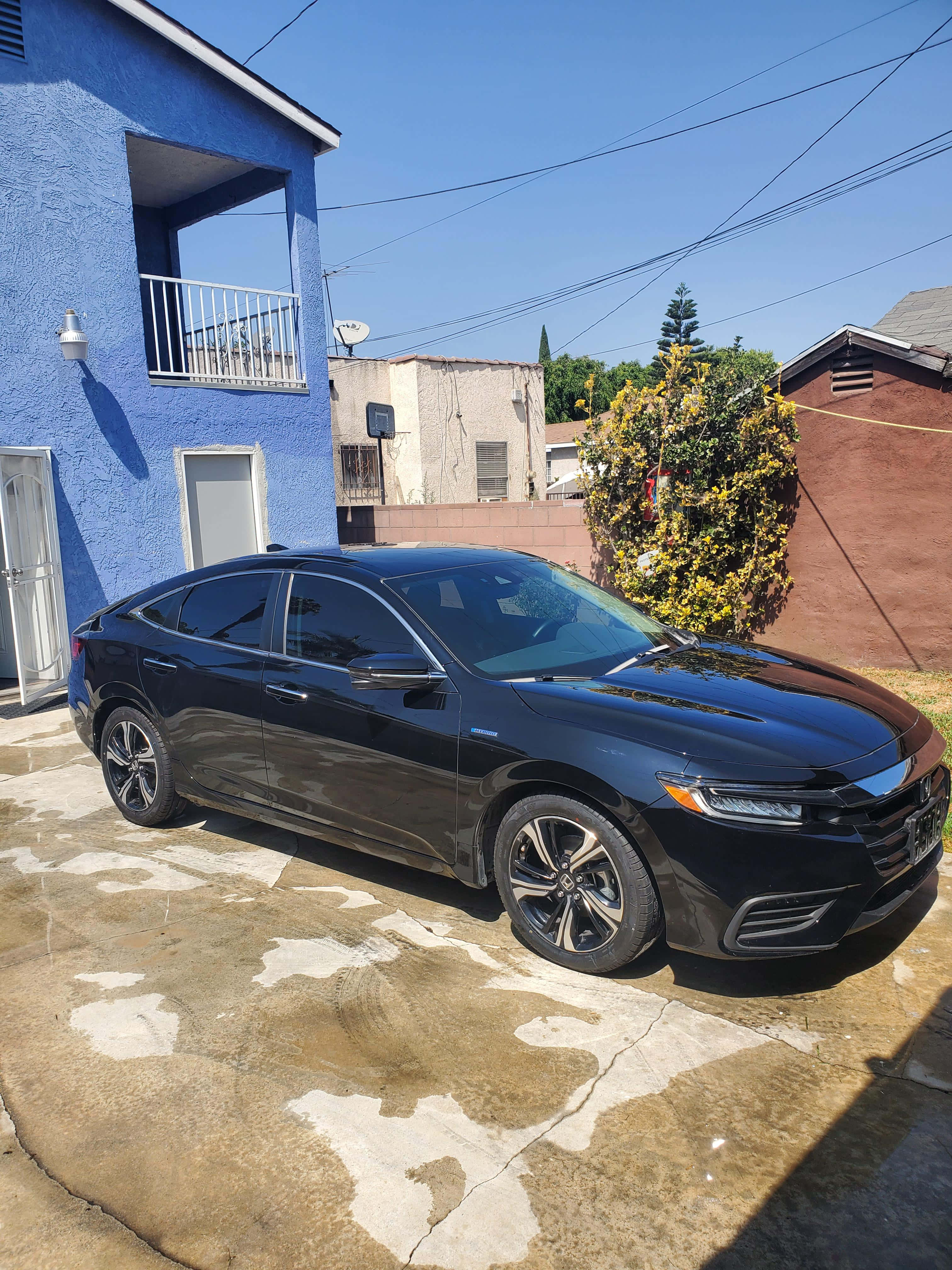 Honda Insight 桌布 壁紙 — 一架時尚黑色轎車停泊在陽光普照的混凝土車道上，旁邊是一棟充滿活力的藍色灰泥房屋，帶有白色陽台。
