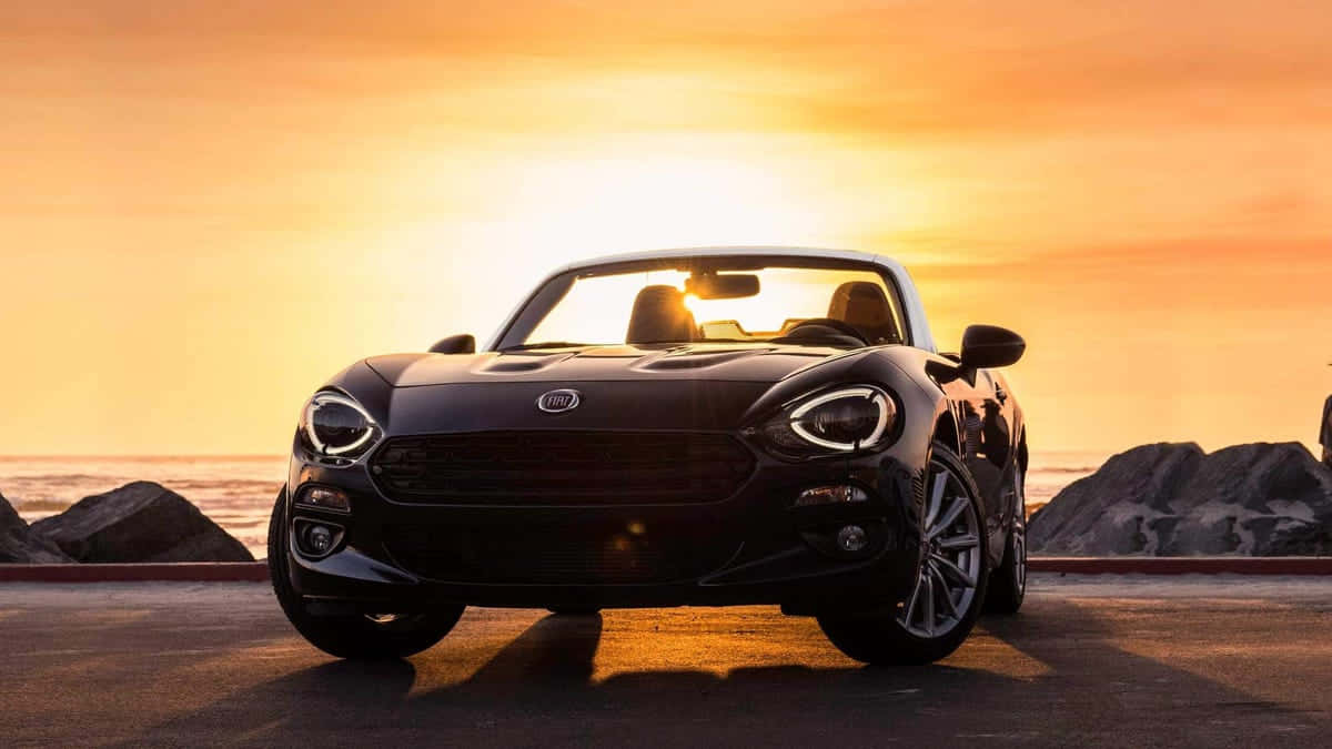 Fiat 124 Spider 桌布 壁紙 — 一架時尚黑色開篷跑車停泊在日落時分的海岸公路上，背景是金色天空和海洋。