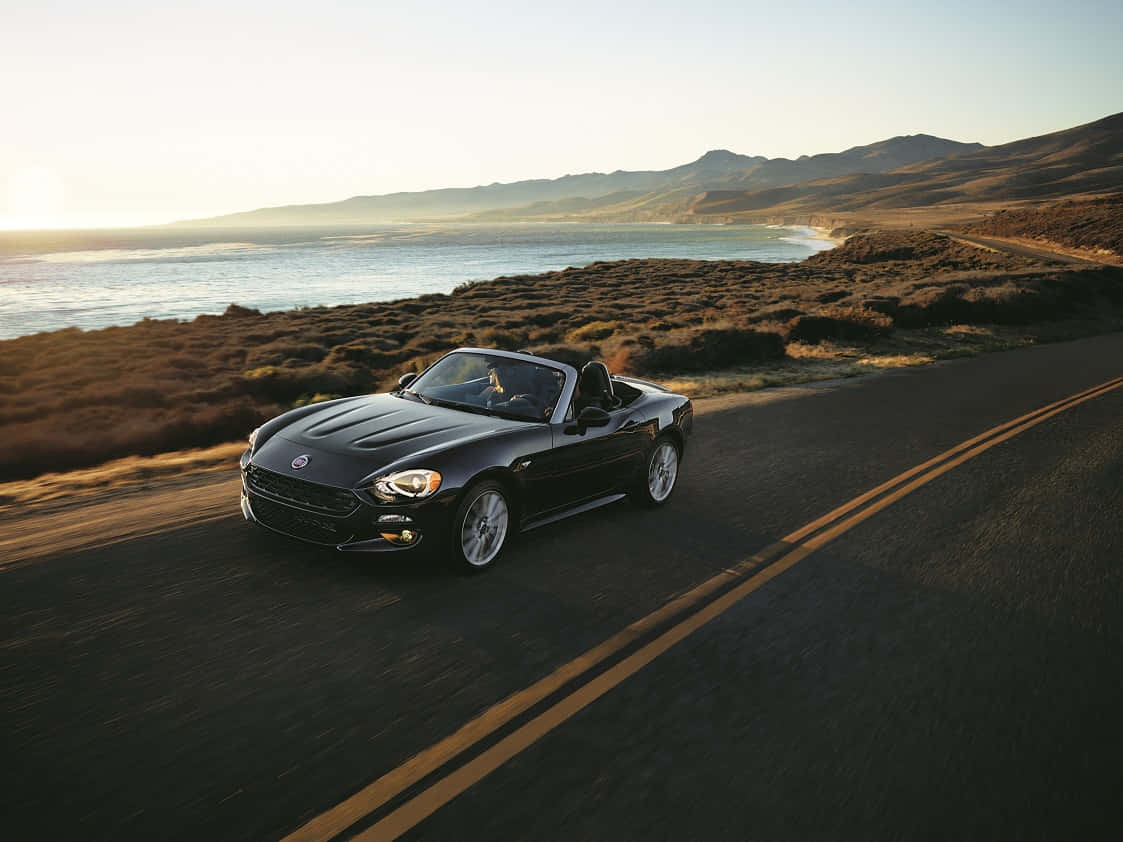 Fiat 124 Spider 桌布 壁紙 — 一架時尚黑色開篷跑車在黃金時段沿著海岸公路行駛，背景是海洋和山丘。