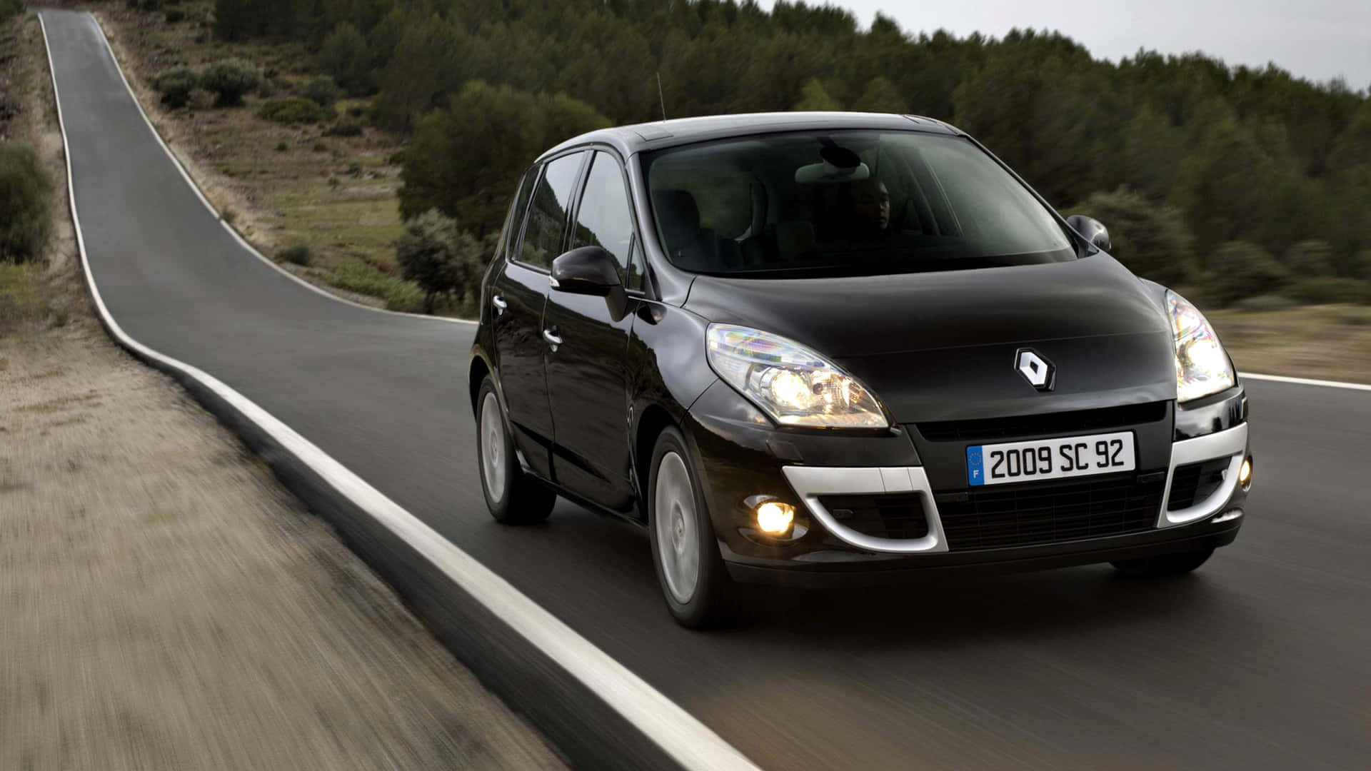 Renault Scenic 桌布 壁紙 — 一輛時尚的黑色雷諾 Scenic 沿著蜿蜒的鄉村道路行駛，兩旁是茂密的綠樹和乾燥的土地。