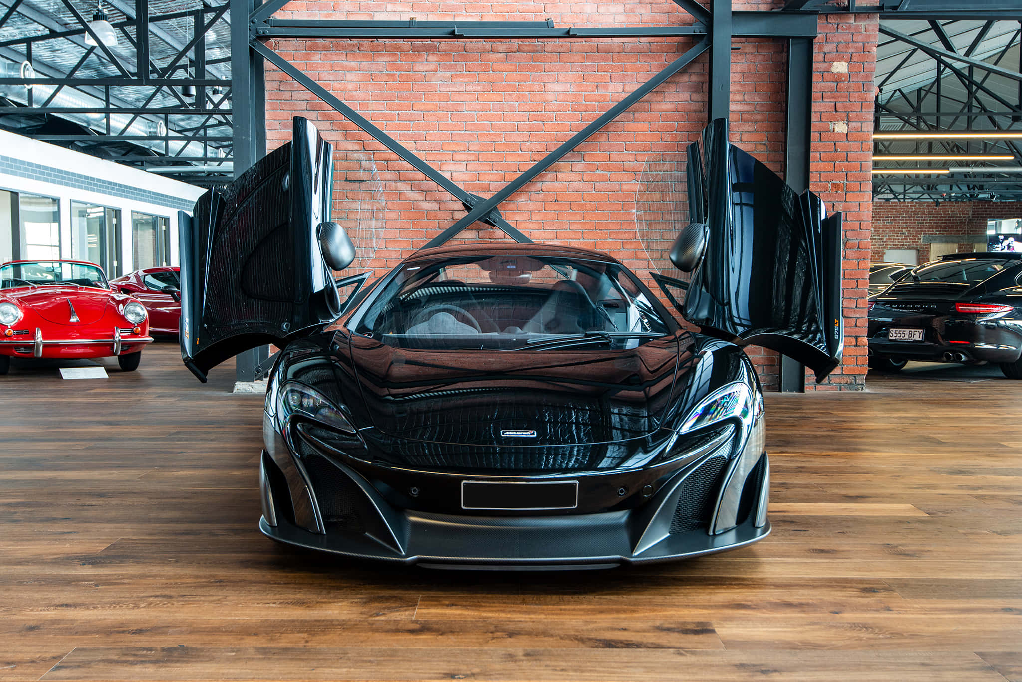 Mclaren 675lt Spider 桌布 壁紙 — 一架時尚的黑色McLaren跑車，鷗翼車門敞開，醒目地停放在寬敞的磚牆車庫中，車庫有木地板，背景還有其他經典汽車。