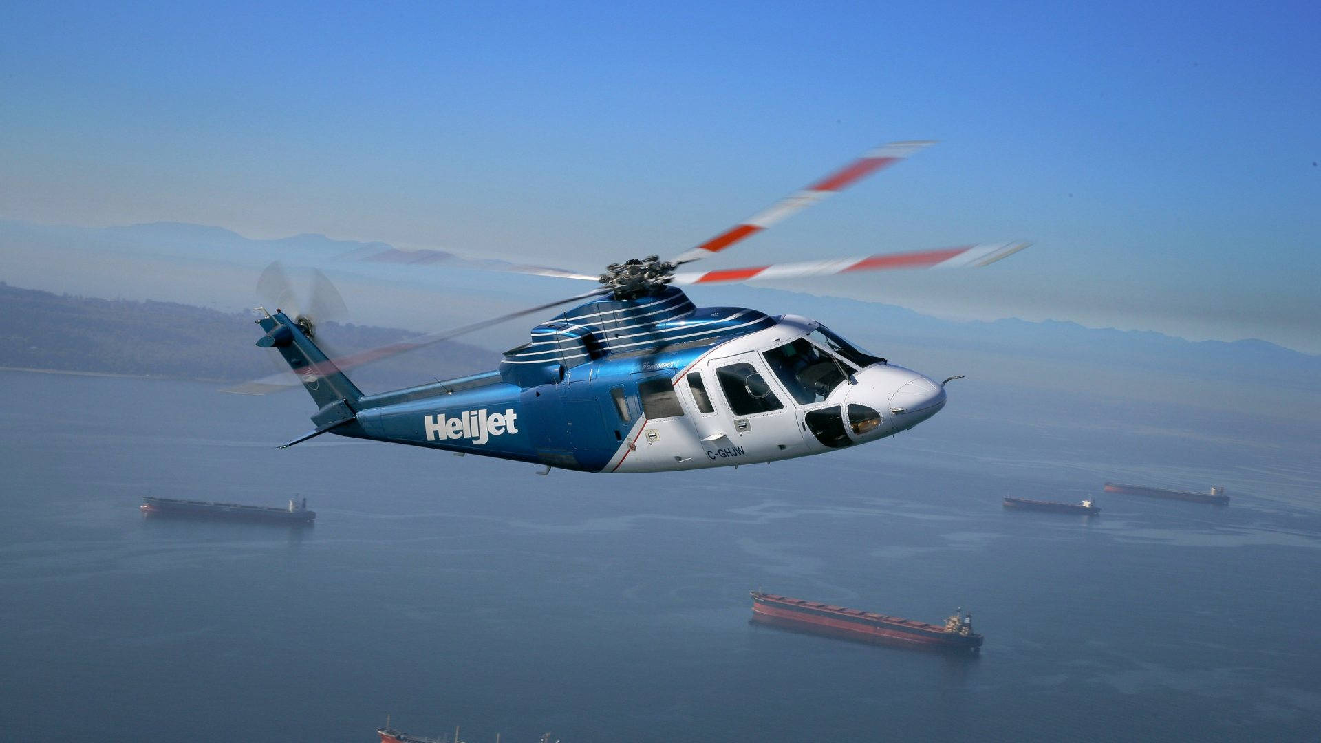 Helijet 直升機 4K 壁紙 — 一架藍白色的Helijet直升機低飛過平靜的海面，下方有貨船，遠處有朦朧的山巒。