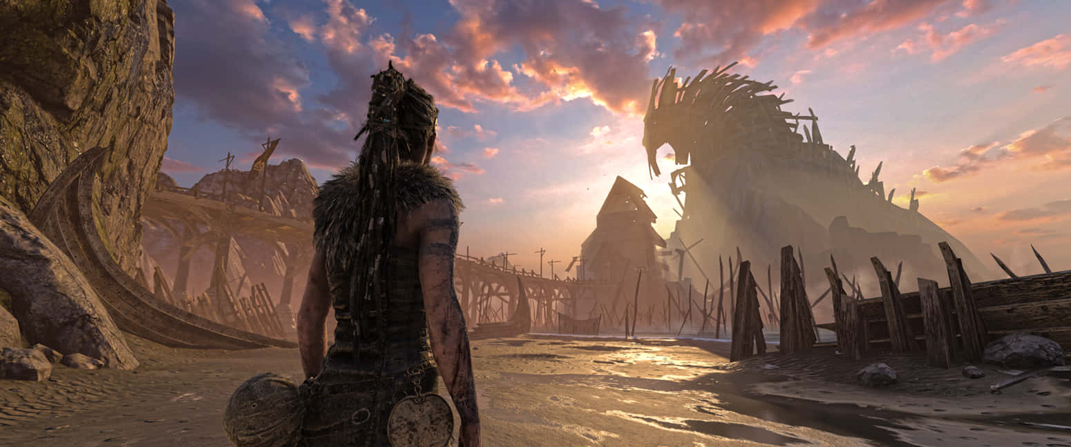 《Hellblade：塞娜的獻祭》3440x1440遊戲場景 壁紙 — 一名戰士站在荒蕪的廢土上，在日落時分，面對著一座巨大的破碎龍雕像，天空如火。