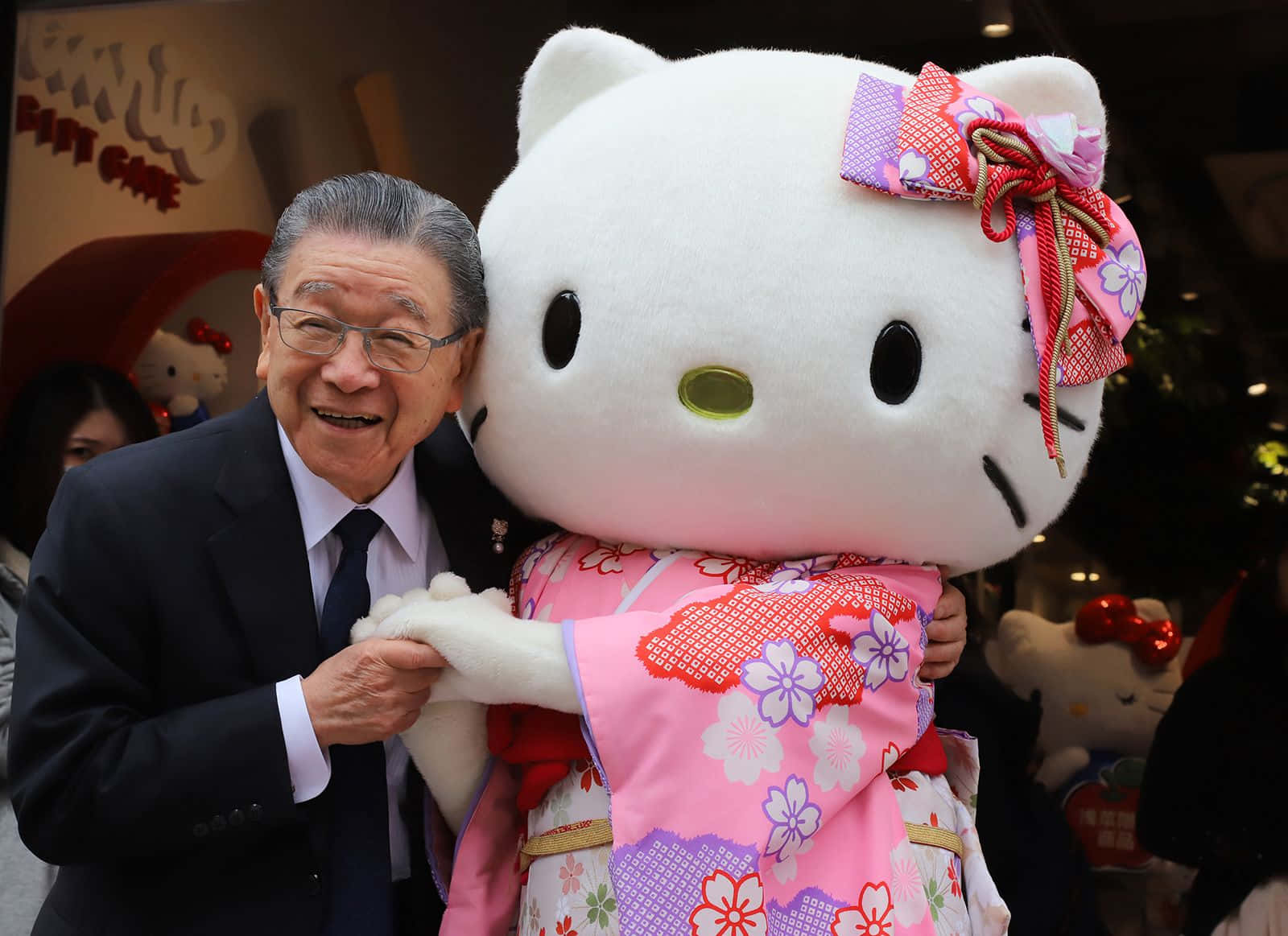 Hello Kitty — 永不褪色的經典貓咪 壁紙 — 一位身穿西裝的長者笑容滿面，擁抱著一個穿著粉紅色花卉和服、繫著圖案蝴蝶結的巨型Hello Kitty吉祥物。