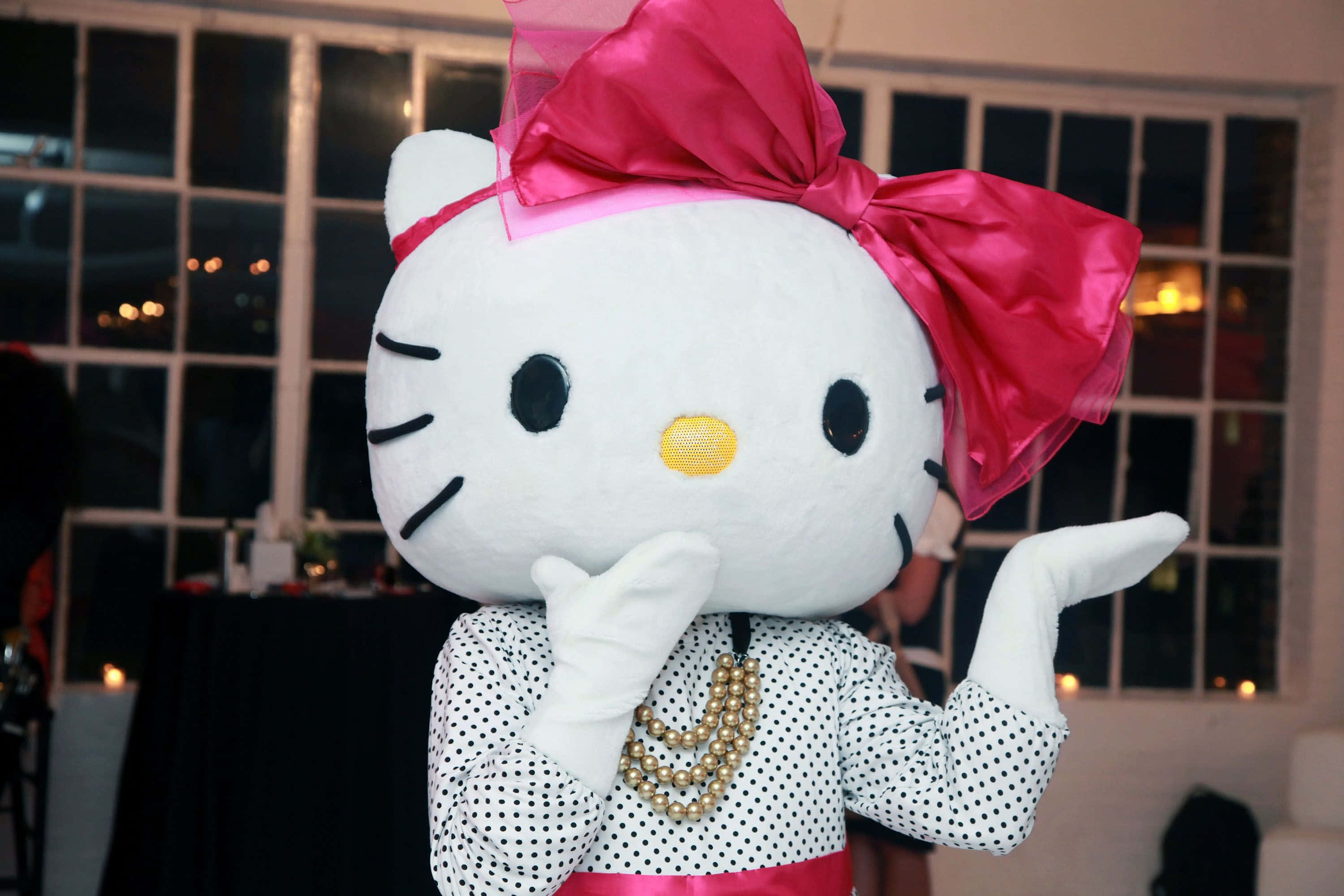Hello Kitty 頭戴粉紅蝴蝶結，周圍環繞粉紫心形 壁紙 — 一個大型Hello Kitty吉祥物穿著波點襯衫和珍珠項鍊，夜晚在窗前單手托腮擺姿勢。