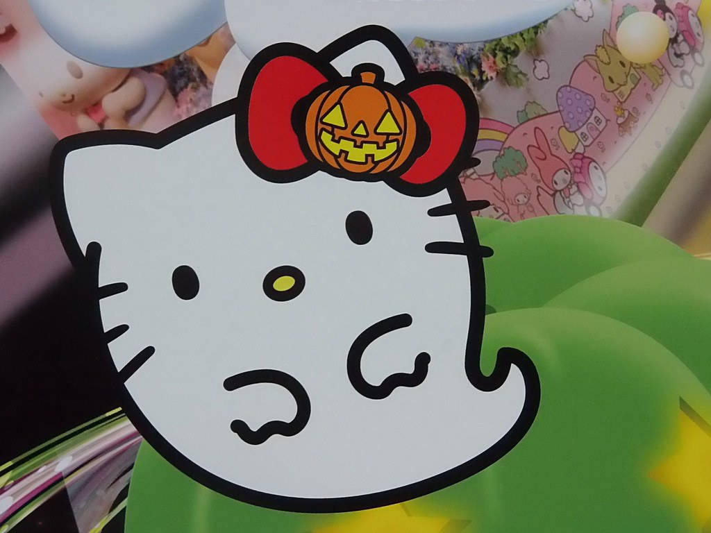 Hello Kitty萬聖節白色幽靈 壁紙 — Hello Kitty 戴著南瓜燈蝴蝶結，在色彩繽紛的卡通背景前開心地微笑。