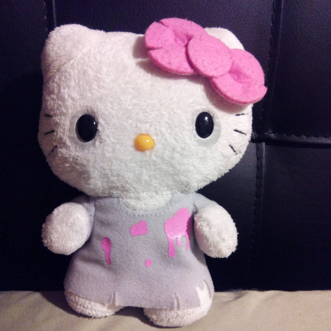 Hello Kitty萬聖節絨毛玩偶 壁紙 — 一隻白色Hello Kitty毛公仔，戴著粉紅色蝴蝶結，灰色裙子上有粉紅色油漆濺點，坐在深色皮革背景前。