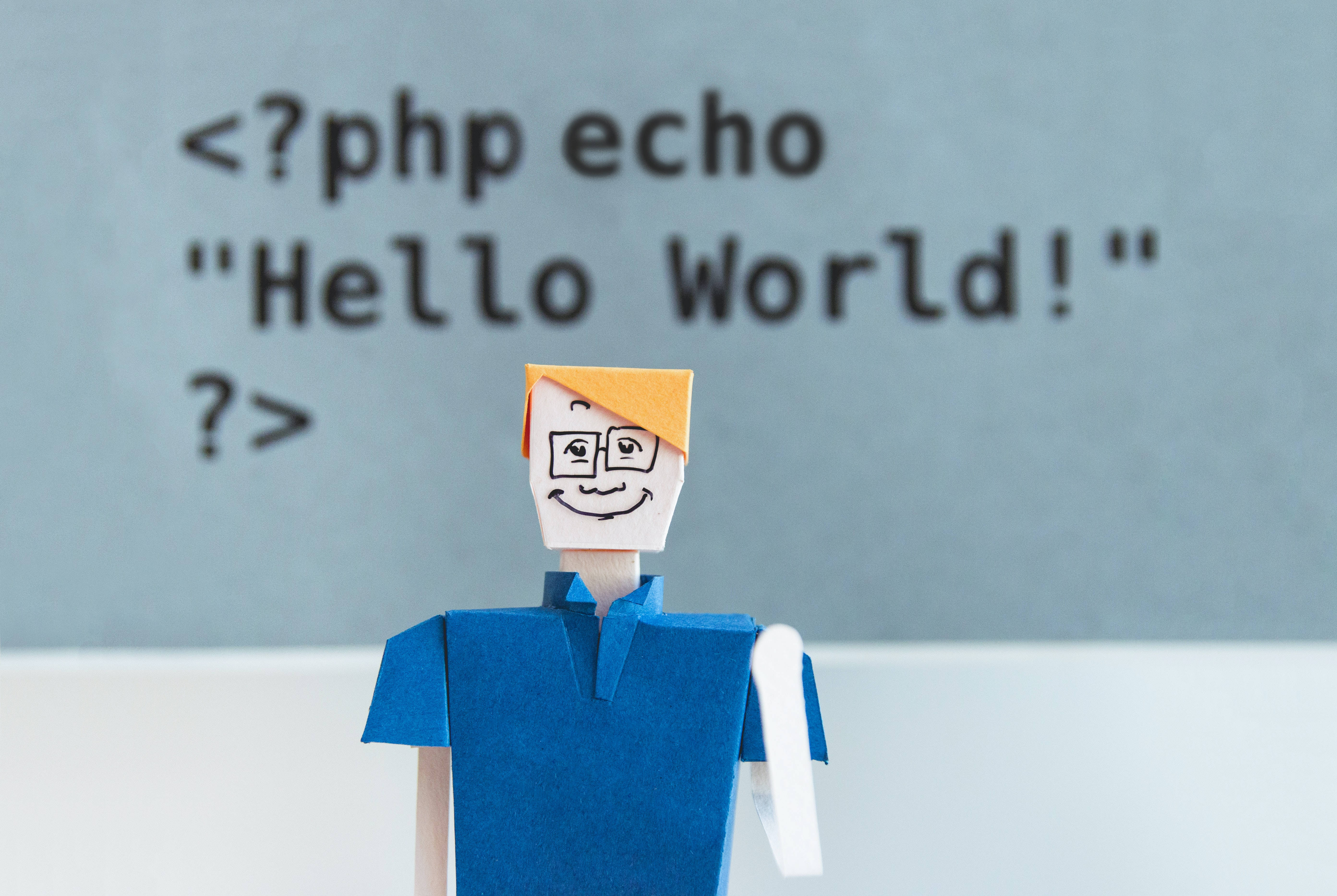「Hello World」藍色紙藝人動畫桌面壁紙 壁紙 — 一個用紙摺成的程式設計師公仔站在灰色牆上模糊的 PHP 'Hello World' 程式碼片段前。