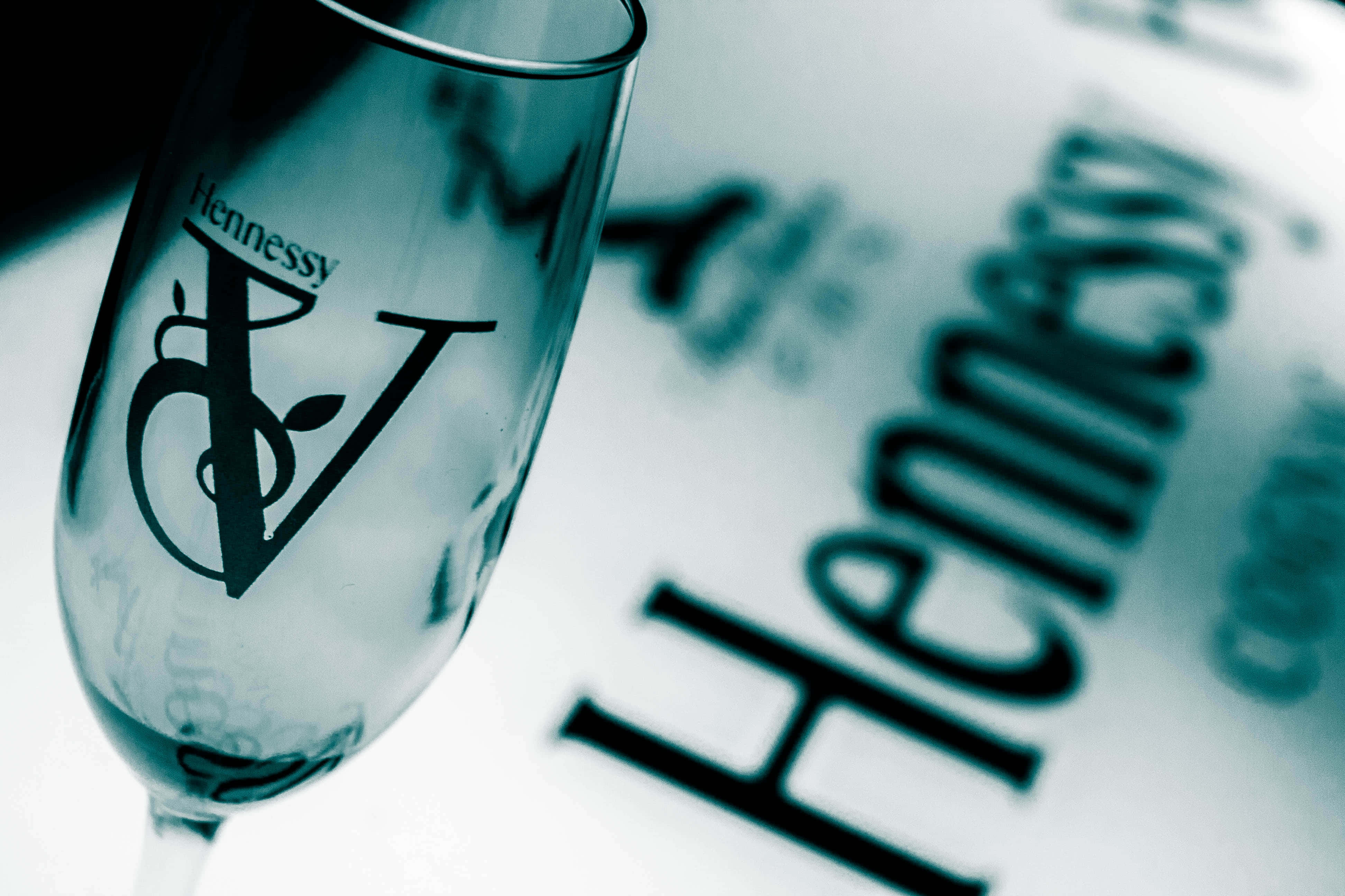 Hennessy 標誌與酒杯 壁紙 — 軒尼詩品牌香檳杯的特寫，放在模糊的軒尼詩標誌表面上，帶有冷藍色調。