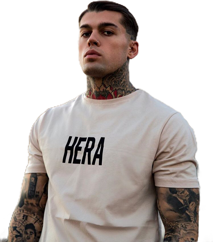 穿HERA T恤的紋身男子 壁紙 — 一位紋身男子身穿印有HERA字樣的米色T恤，站在黑色背景前。
