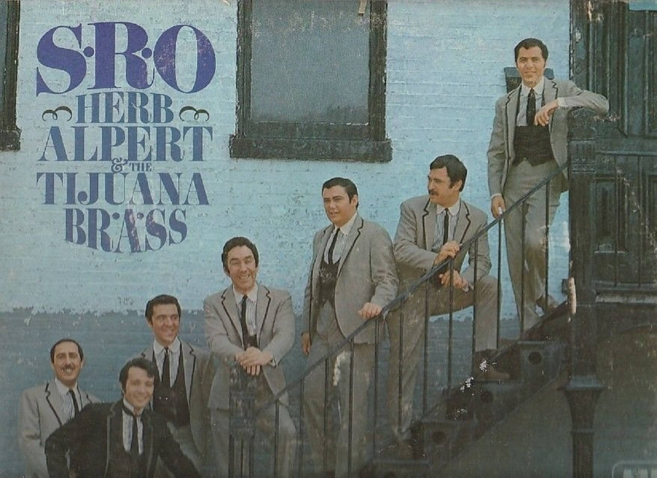 Herb Alpert And The Tijuana Brass S.R.O. 專輯 壁紙 — 七名身穿灰色西裝的男子在一個風化的戶外樓梯上擺姿勢，旁邊是一面褪色的藍色牆壁，上面有'Herb Alpert & The Tijuana Brass'的字樣。