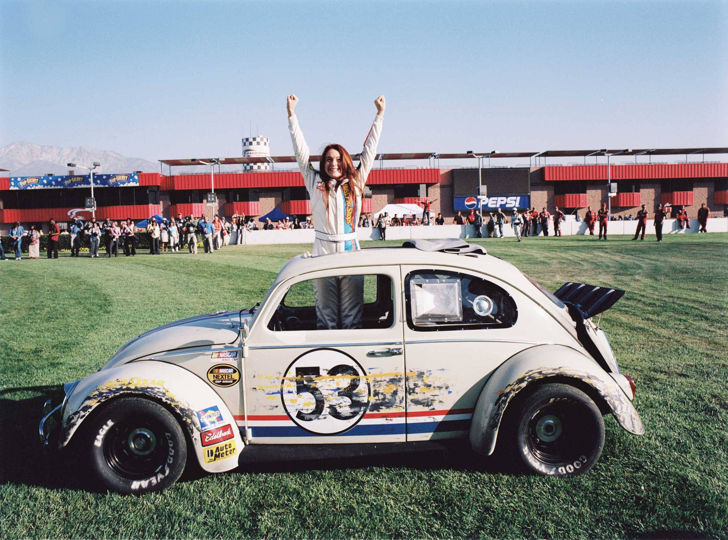 《Herbie：Fully Loaded》Lindsay Lohan高舉雙臂 壁紙 — 一位快樂的女賽車手在她白色的#53改裝福士甲蟲車頂上慶祝，背景是藍天和草地賽道。