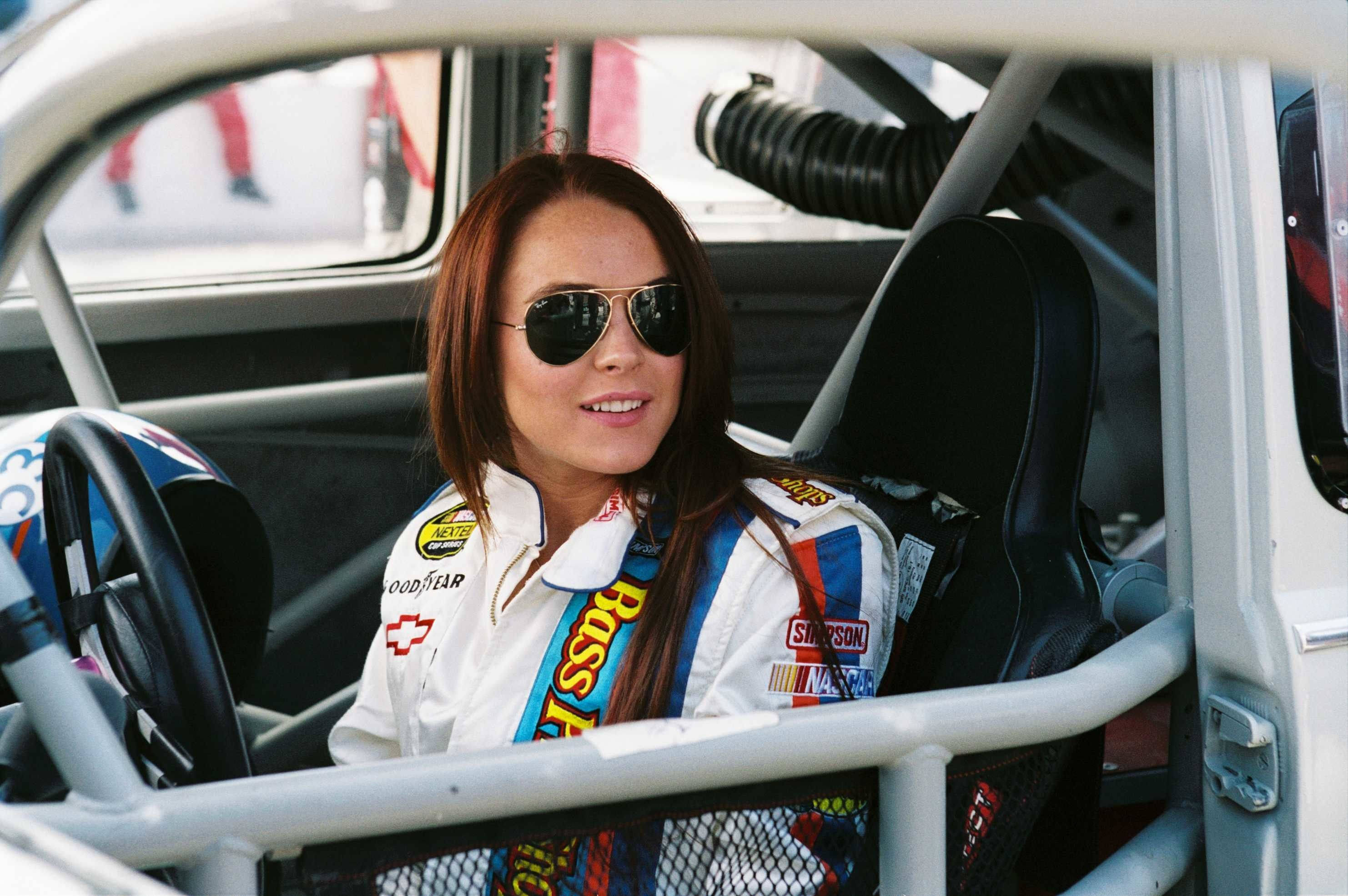 《Herbie：Fully Loaded》琳賽·蘿涵駕駛座壁紙 壁紙 — 一位微笑的女賽車手，身穿白色贊助商賽車服，自信地坐在改裝房車的駕駛艙內。