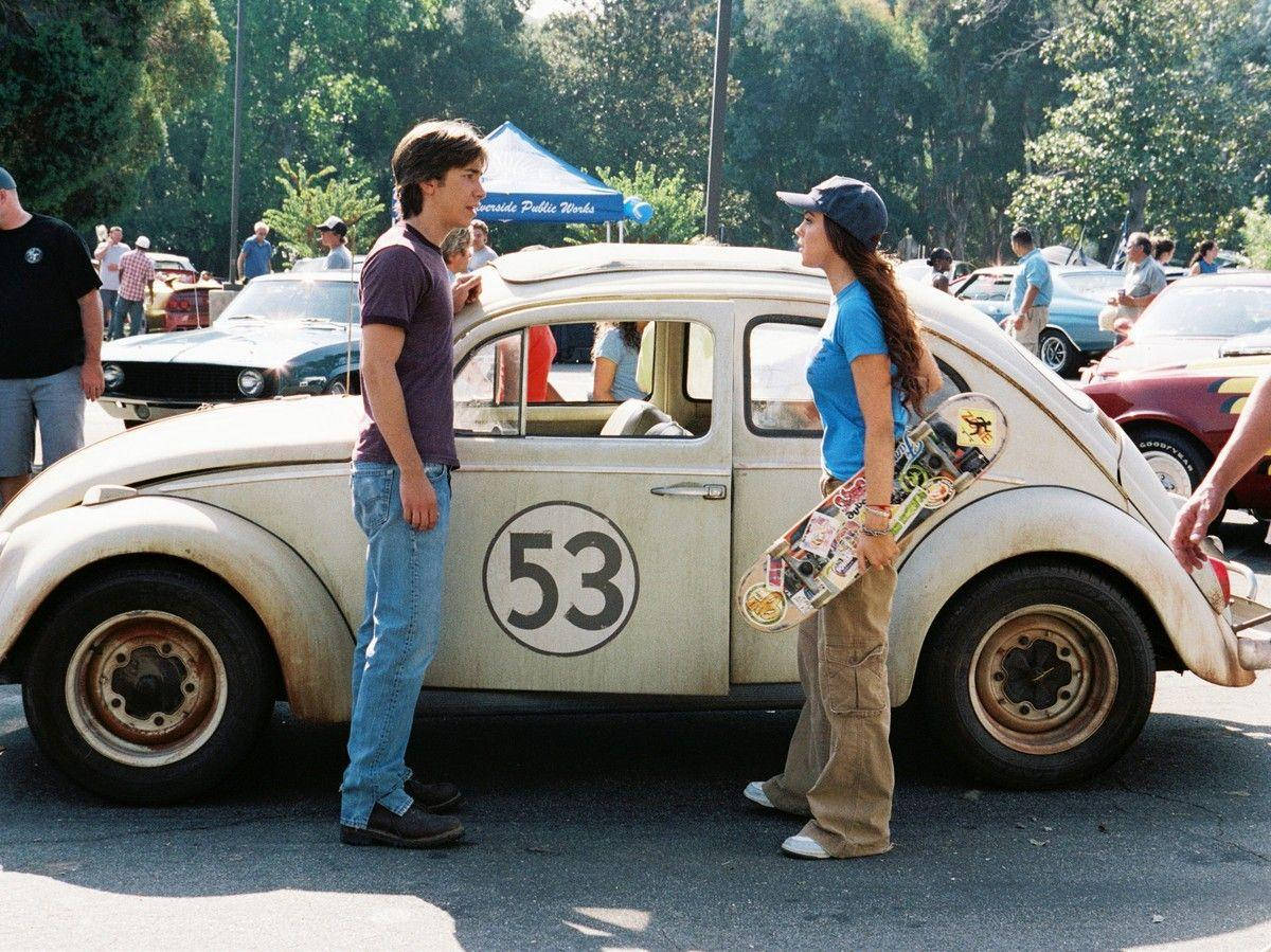 《Herbie Fully Loaded》情侶與滑板 壁紙 — 一名年輕男子和女子在一輛白色復古福士甲蟲車（車牌號碼53）旁邊交談，該車停泊在陽光明媚的戶外車展上。