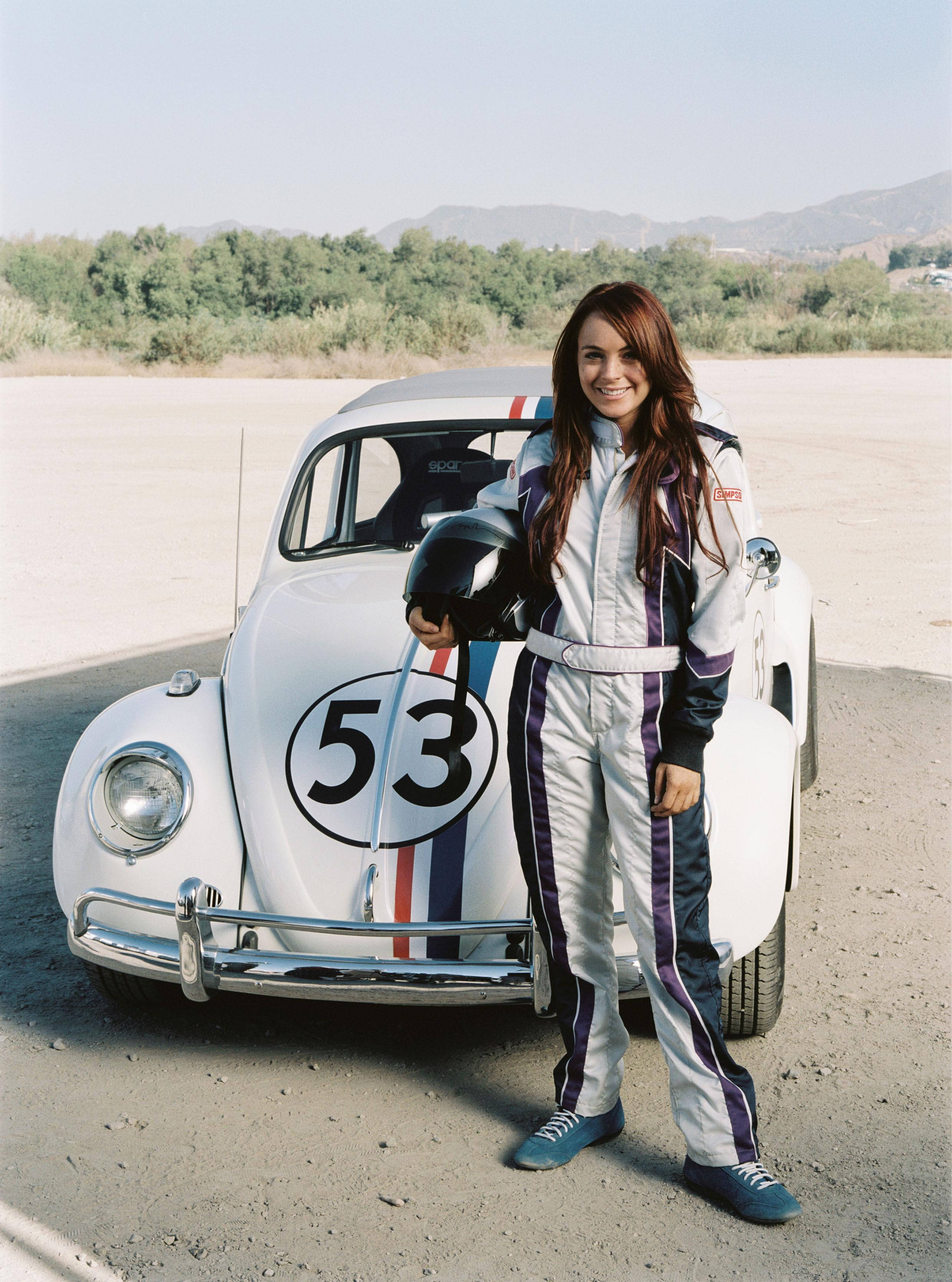 《Herbie Fully Loaded》琳賽·蘿涵與Herbie 壁紙 — 一位微笑的女性穿著賽車服，驕傲地站在一架白色復古賽車旁，車上標有53號，背景是陽光明媚的飛機跑道。