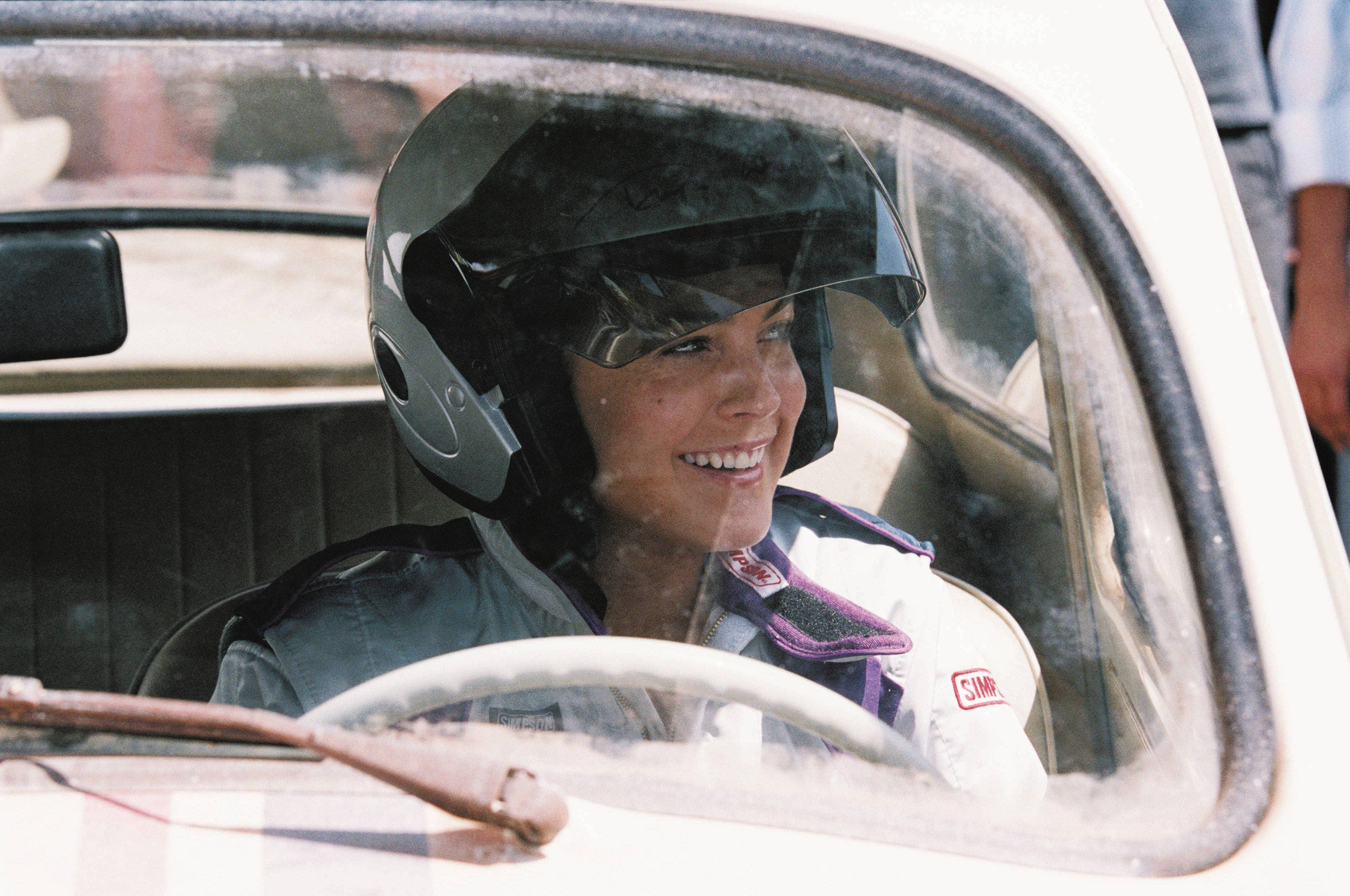 《Herbie：Fully Loaded》微笑的琳賽·蘿涵壁紙 壁紙 — 一位微笑的女性戴著賽車頭盔和賽車服，坐在一輛復古賽車內，愉快地望向窗外。