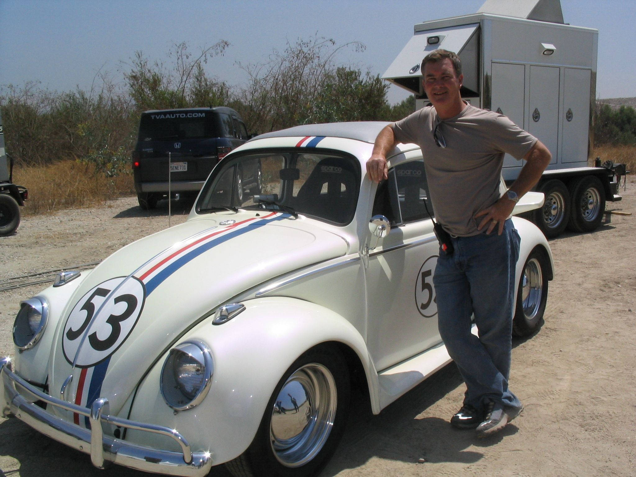 《Herbie：Fully Loaded》與男子同框壁紙 壁紙 — 一名男子在白色復古賽車旁微笑，賽車帶有紅藍條紋和53號，停在塵土飛揚的地形上。