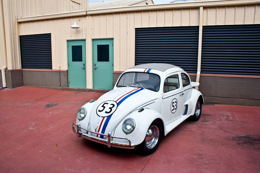 《Herbie：Fully Loaded》Herbie停靠於建築旁 壁紙 — 一輛帶有紅藍條紋和53號的白色復古賽車停泊在紅色混凝土場地上，前面是帶有綠色門的米色建築。