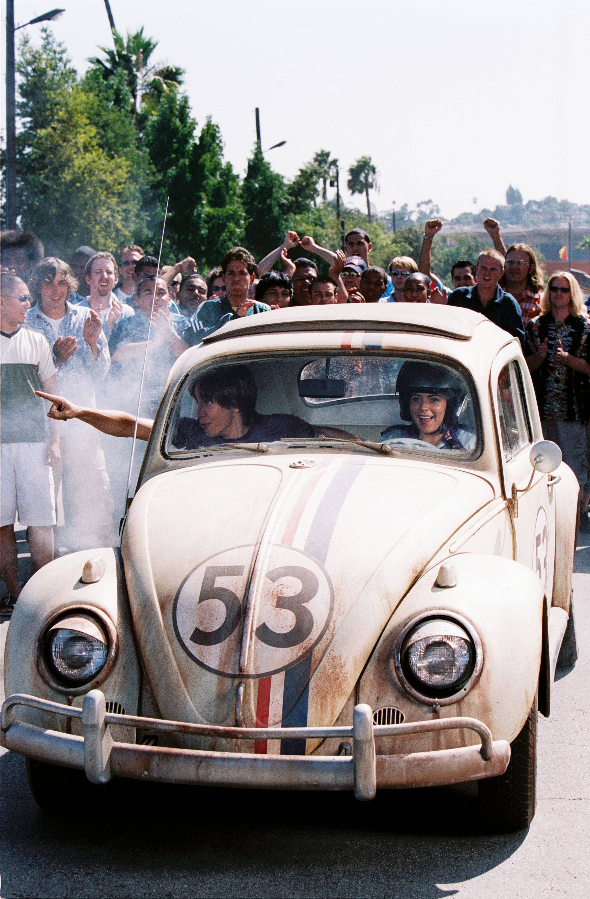 《Herbie：Fully Loaded》情侶駕駛群眾壁紙 壁紙 — 一輛復古白色福士甲蟲車（編號53）在陽光明媚的街道上奔馳，受到熱鬧人群的歡呼。