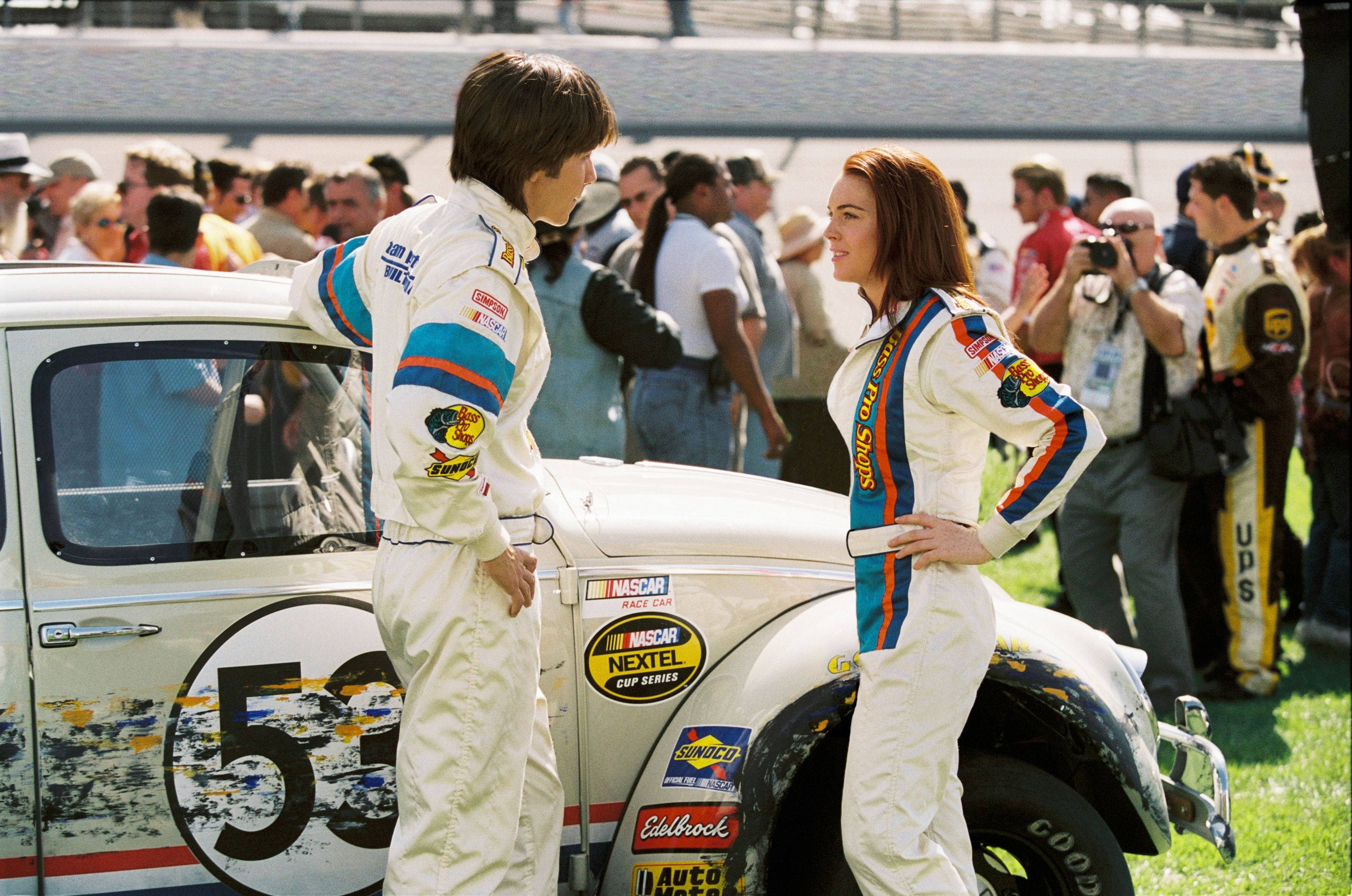 《Herbie：Fully Loaded》情侶同框壁紙 壁紙 — 兩名身穿白色賽車服的賽車手靠在一輛編號53的復古NASCAR賽車上，觀眾在後面觀看。