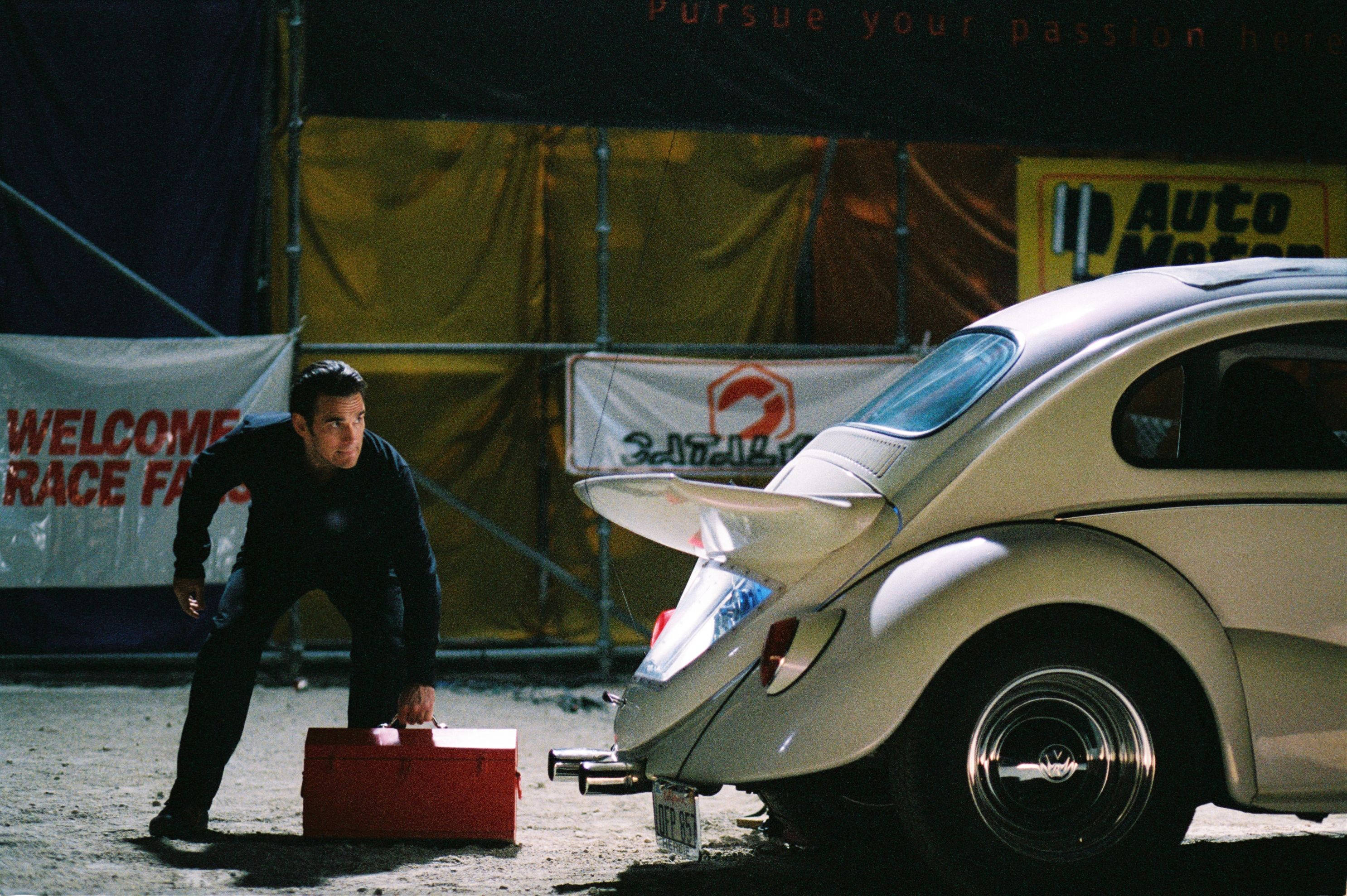 《Herbie：Fully Loaded》馬特·狄龍壁紙 壁紙 — 一名穿黑色衣服的男子在賽道上蹲在一個紅色工具箱旁邊，旁邊是一輛改裝過的白色福士甲蟲車。