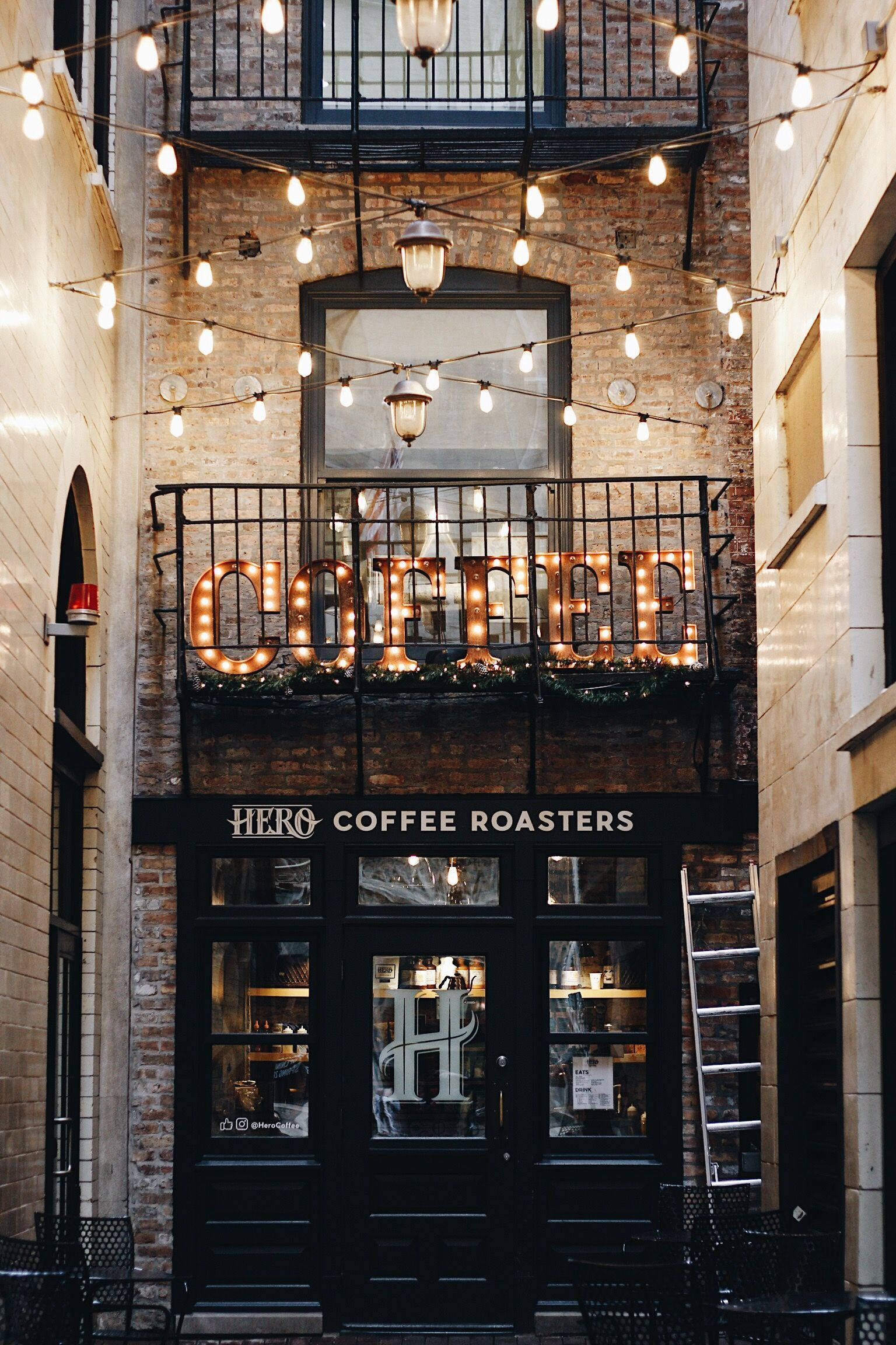 芝加哥 HERO 咖啡館 壁紙 — 一間舒適的城市咖啡店，在黃昏時分，磚砌建築上有溫暖的串燈和發光的「COFFEE」標誌。