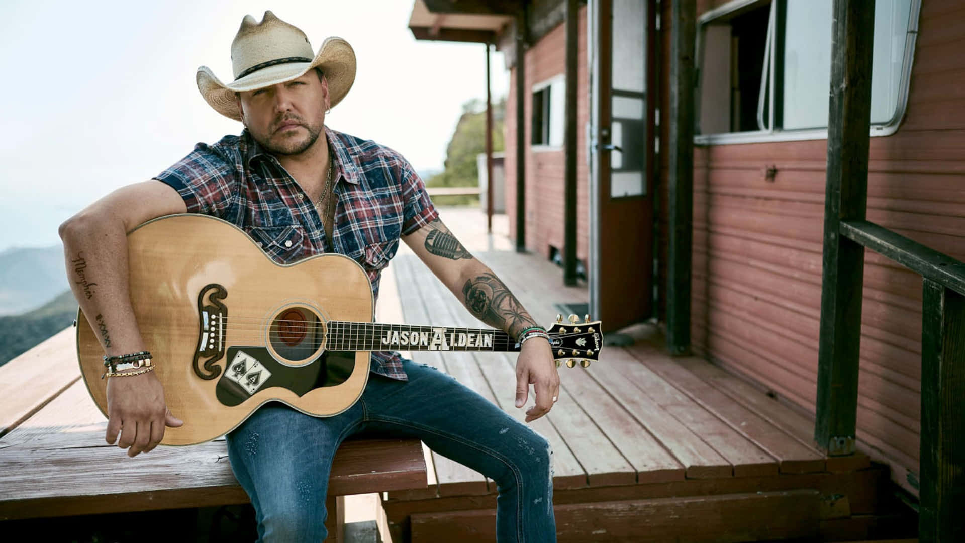  壁紙 — 鄉村歌手Jason Aldean坐在木製門廊上，手持一把原聲結他，手臂上的紋身清晰可見。