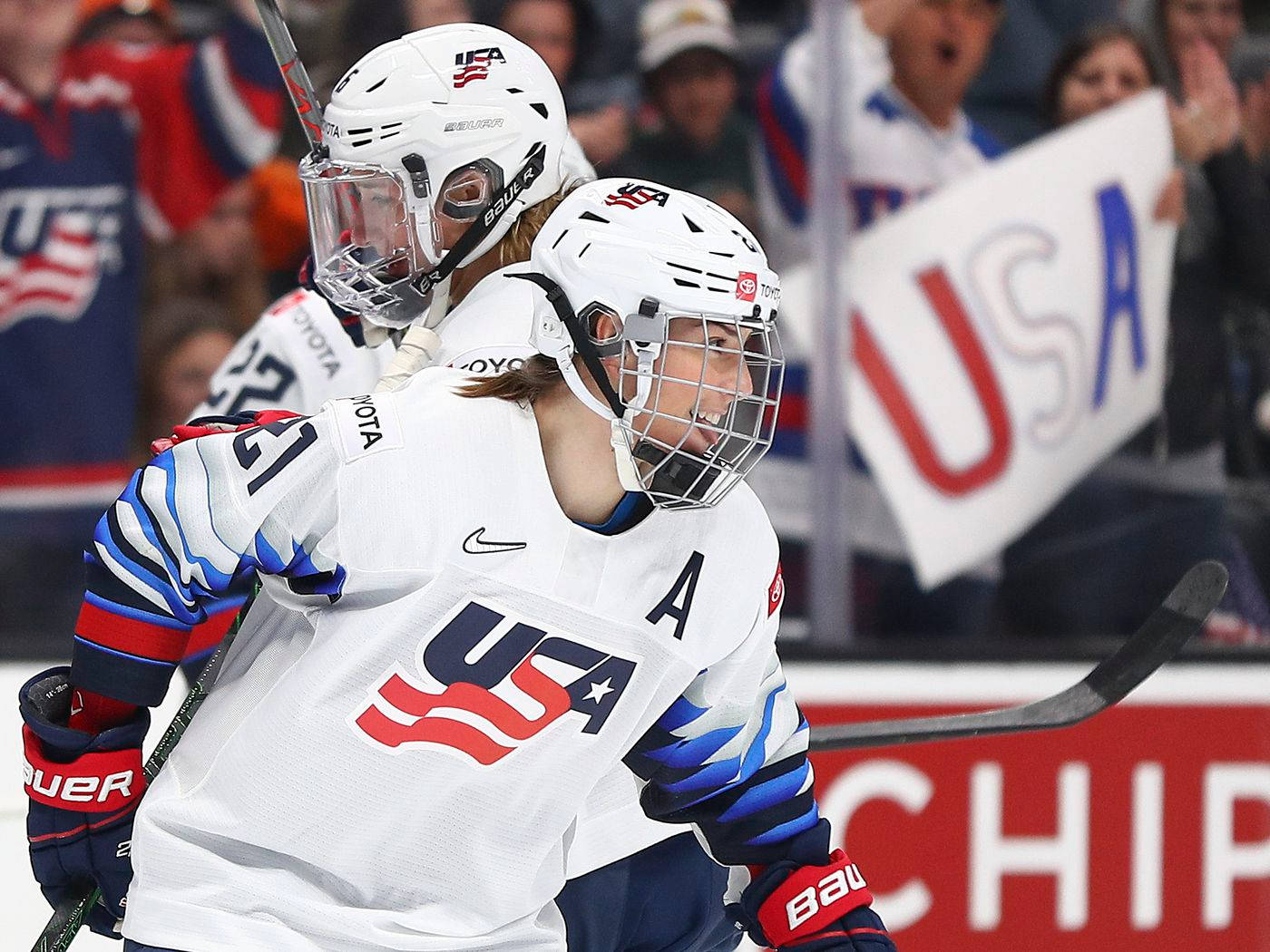 Hilary Knight 世界級運動員 壁紙 — 兩名身穿白色球衣的美國曲棍球運動員在冰上慶祝入球，身後有球迷歡呼。
