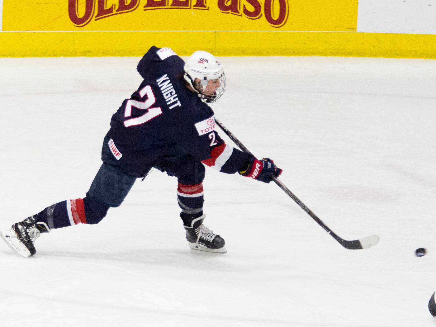 Hilary Knight，冰上冠軍 壁紙 — 一名身穿深藍色球衣、號碼21的冰球運動員在明亮的場館燈光下，在冰上激烈前傾，在冰球附近控球。