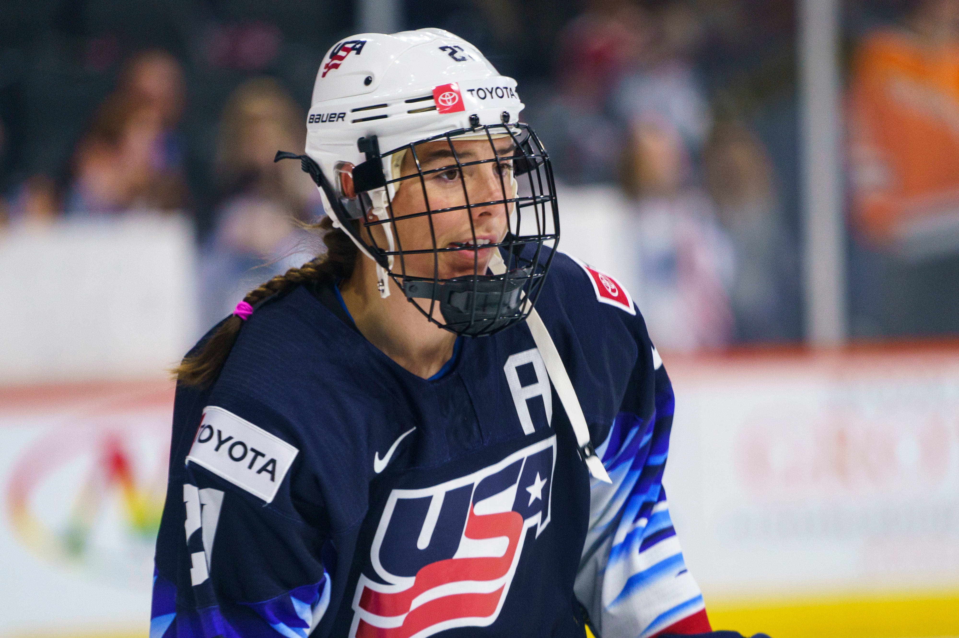 Hilary Knight 美國冰球前鋒 壁紙 — 一位專注的女冰球運動員，身穿美國隊球衣和頭盔，在明亮的場館燈光下於比賽中凝視前方。