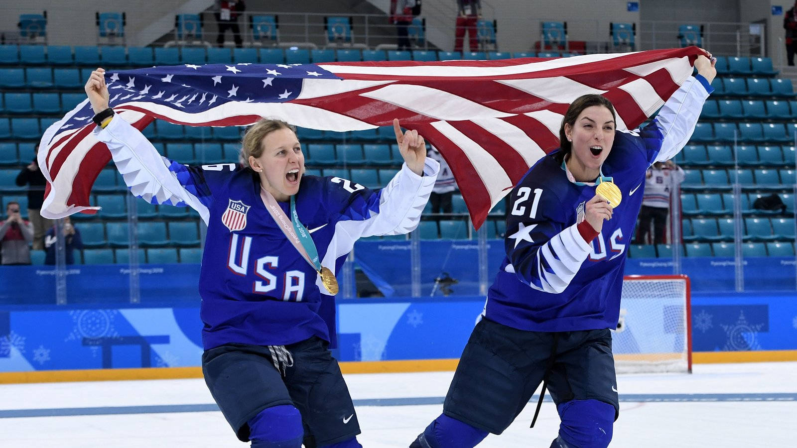 Hilary Knight 與 Kendall Coyne 2018 冬奧金牌 壁紙 — 兩名歡欣鼓舞的美國冰球運動員在冰場上手持大型美國國旗和金牌慶祝。