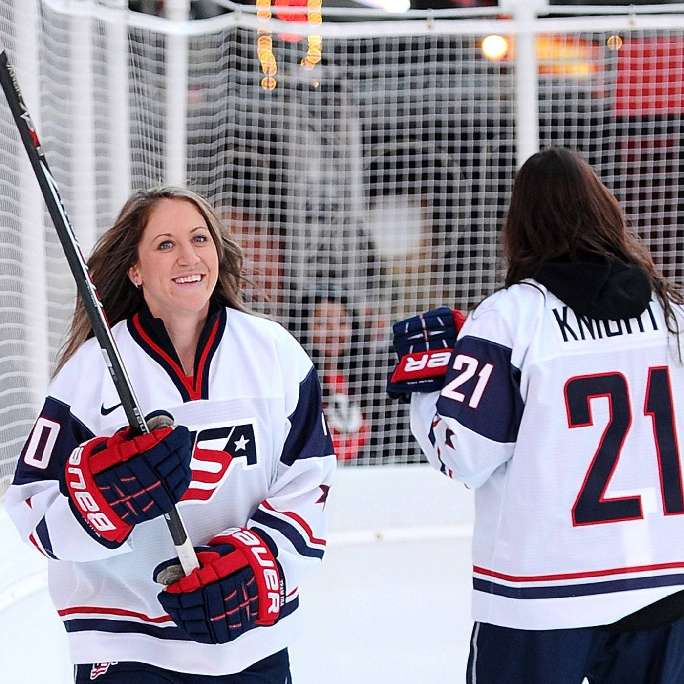 Hilary Knight 與隊友 Meghan Duggan 壁紙 — 兩名身穿美國球衣、面帶微笑的女子冰上曲棍球運動員在冰場上拿著球桿和手套分享歡樂時刻。