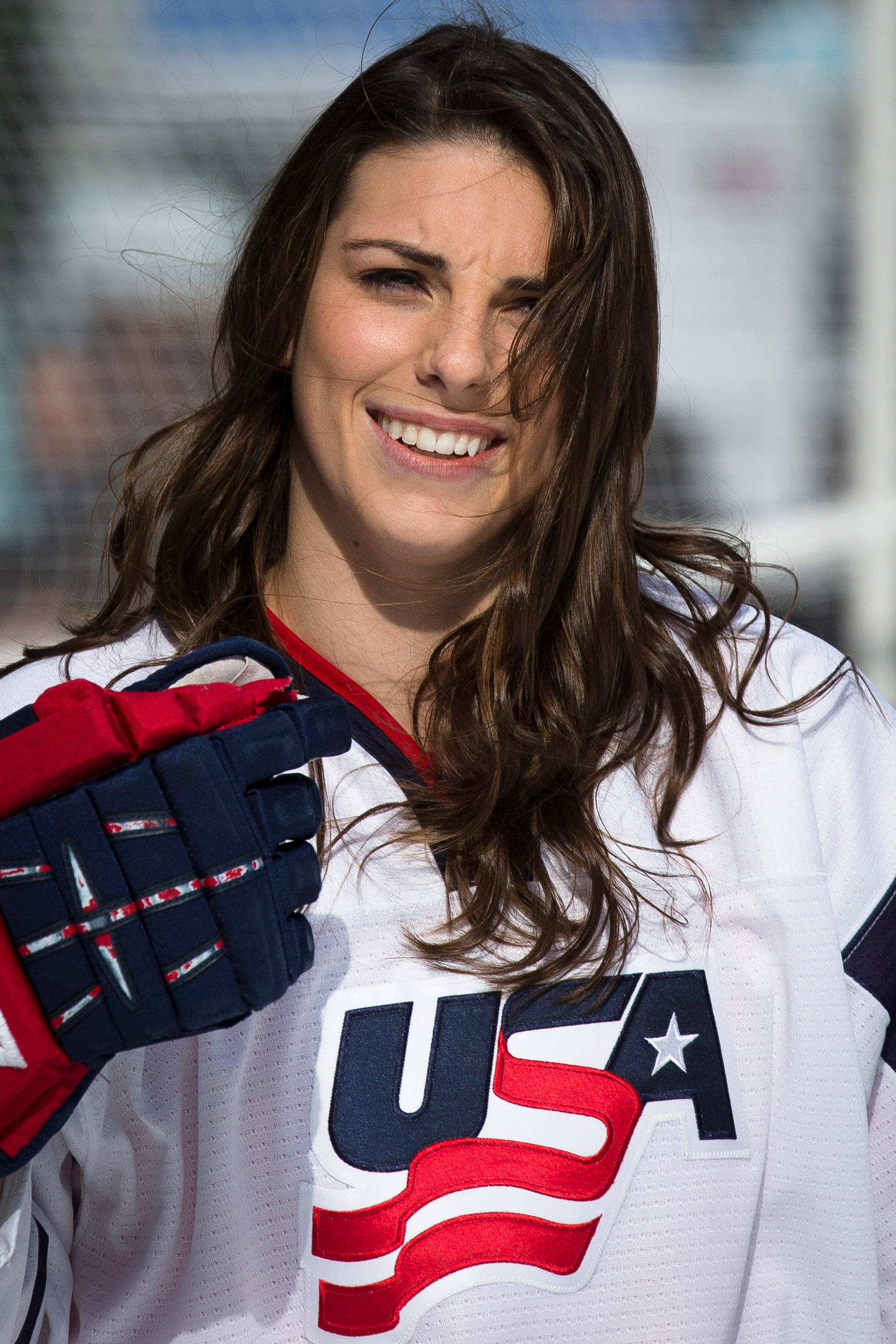 Hilary Knight 近距離肖像照 壁紙 — 一位身穿美國球衣、面帶微笑的女子冰上曲棍球運動員在明亮的戶外背景下舉起手套。