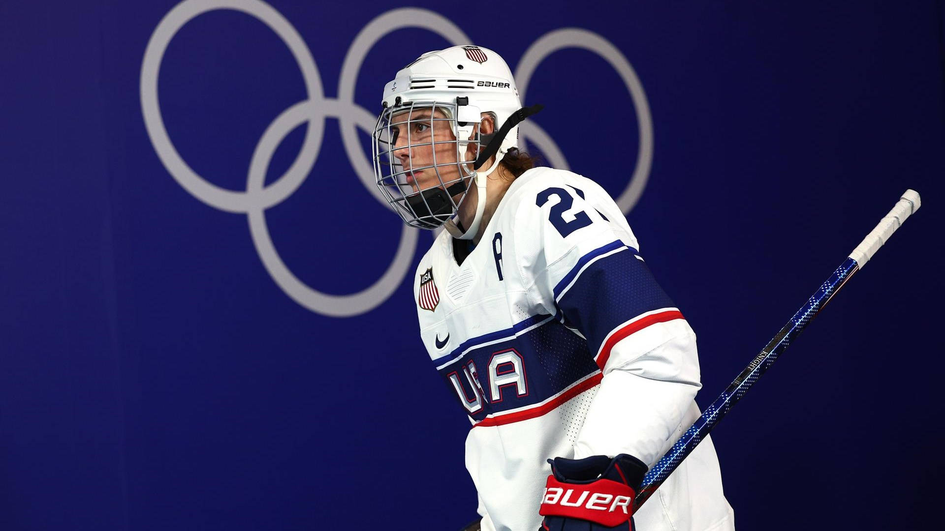 Hilary Knight 頂尖奧運運動員 壁紙 — 一名專注的美國冰球運動員身穿白色裝備，在帶有奧運五環的藍色背景下持球棍。