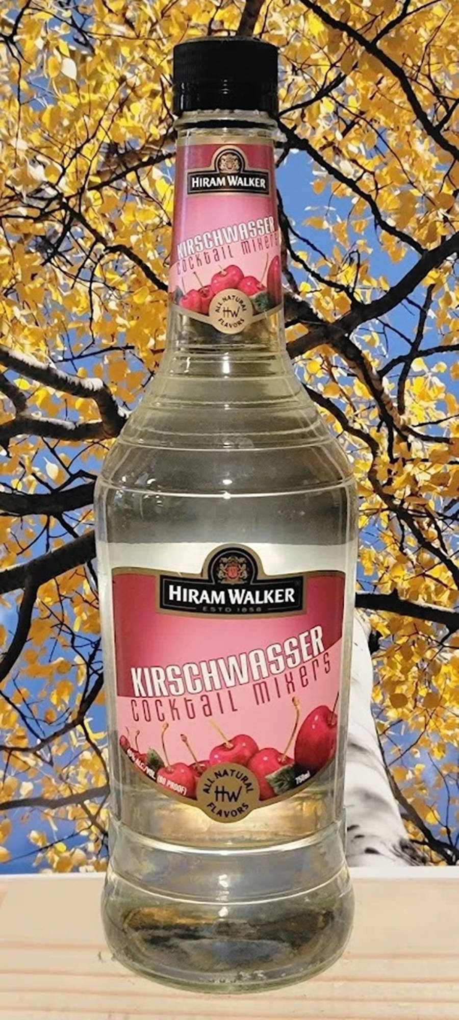 Hiram Walker 樱桃利口酒調酒配料攝影 壁紙 — 一瓶透明的Hiram Walker Kirschwasser雞尾酒調和劑，以金色秋葉和藍天為背景。