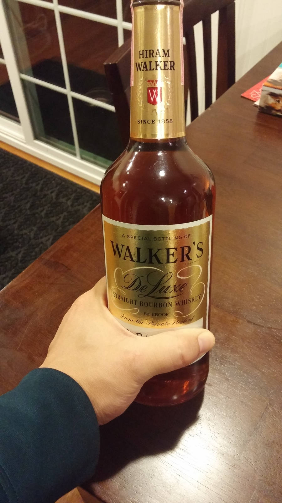 Hiram Walker 高級直火波本威士忌瓶 壁紙 — 一隻手喺窗邊嘅木枱上拎住一支Walker's Deluxe波本威士忌酒樽。