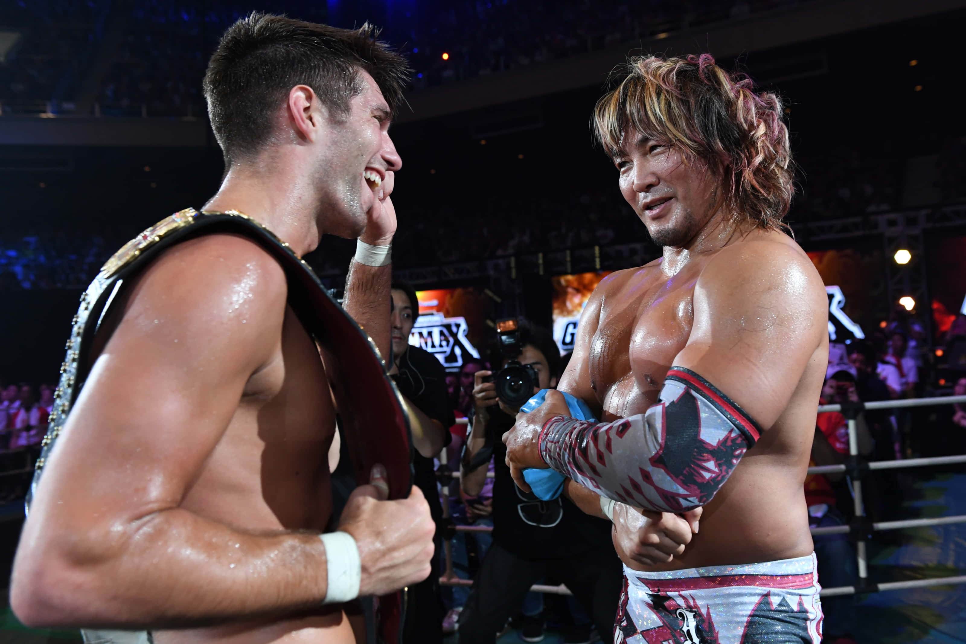 Hiroshi Tanahashi 與 Zack Sabre Jr 壁紙 — 兩名汗流浹背的職業摔角手在燈光明亮的擂台上咧嘴笑著並互相打手勢，背景是歡呼的粉絲。