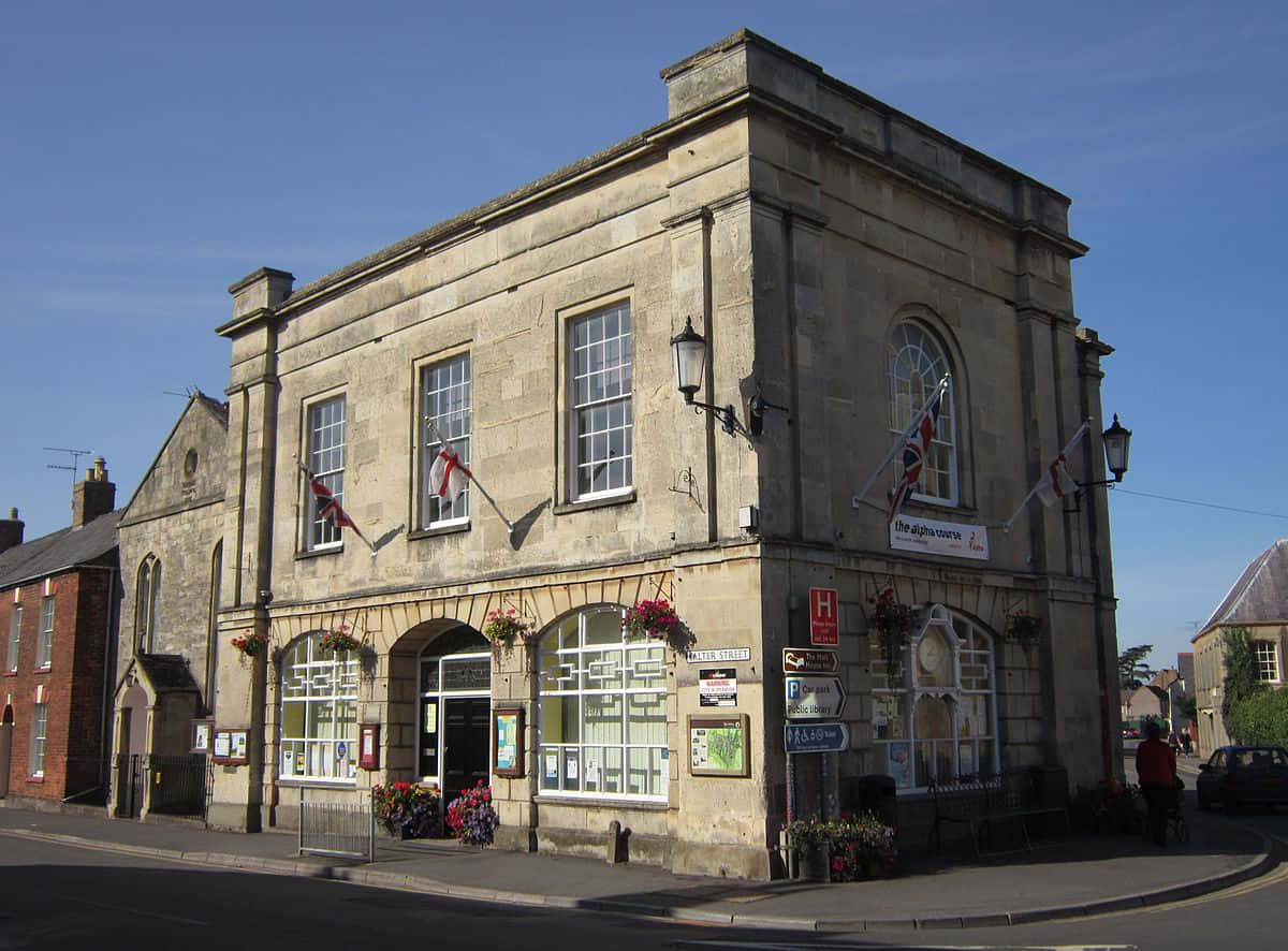 Gloucester United Kingdom 桌布 壁紙 — 一座歷史悠久的石建築，擁有拱形窗戶和懸掛的花籃，矗立在晴朗藍天下的街角。